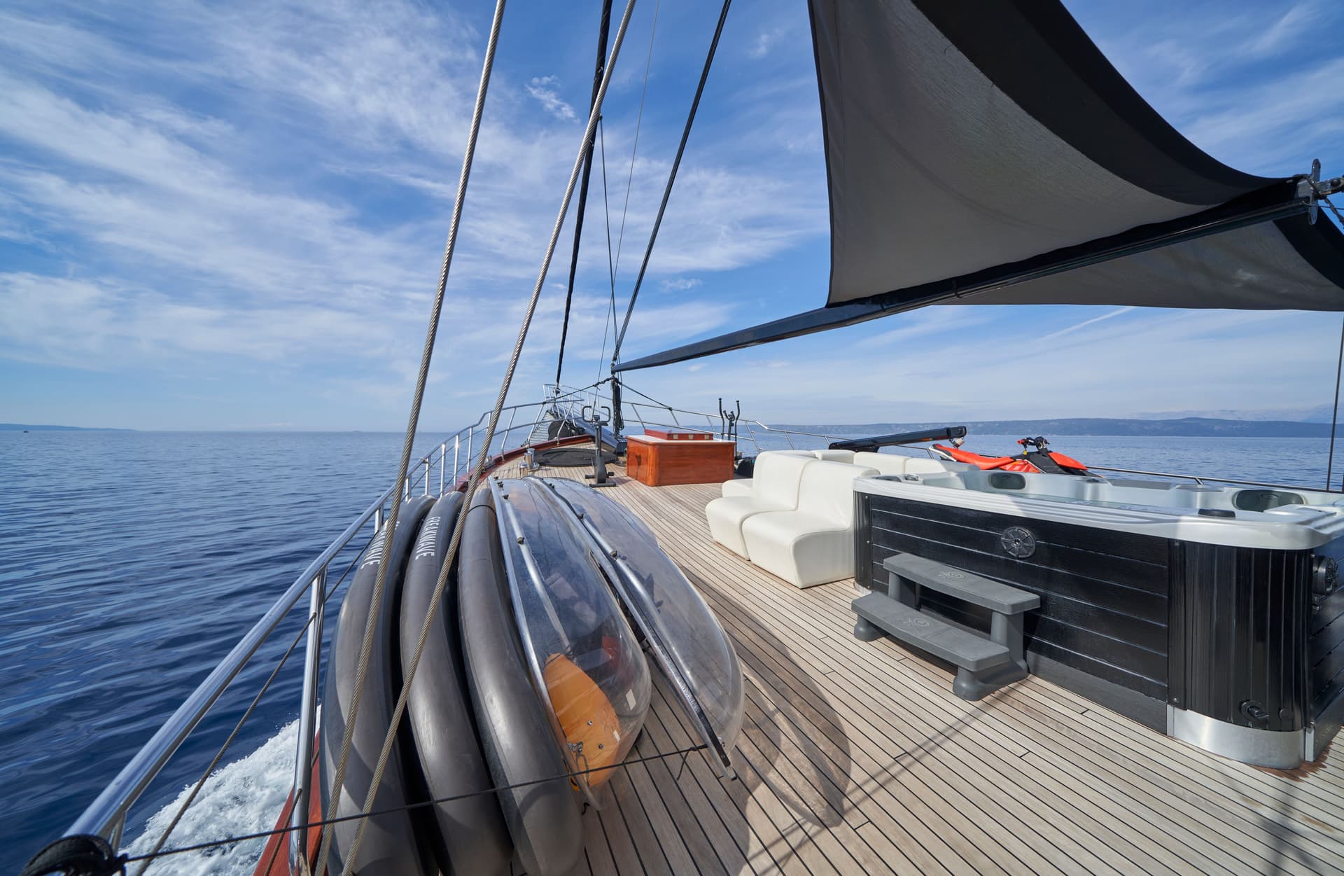 Yacht deck with teak flooring, hot tub, white seating, and kayaks on blue sea.
