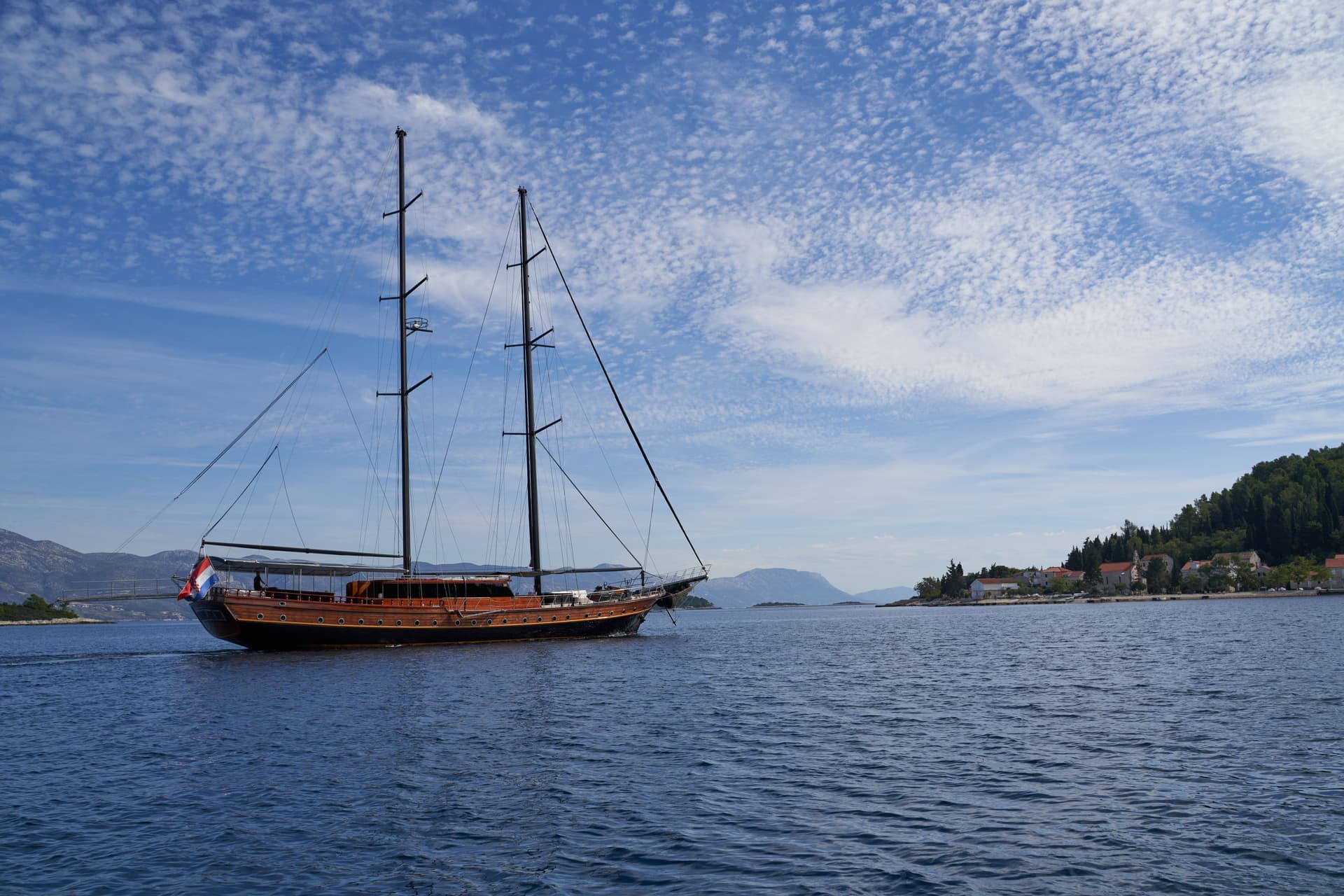 Wooden sailboat with Croatian flag on blue water near a forested Croatian coastline.