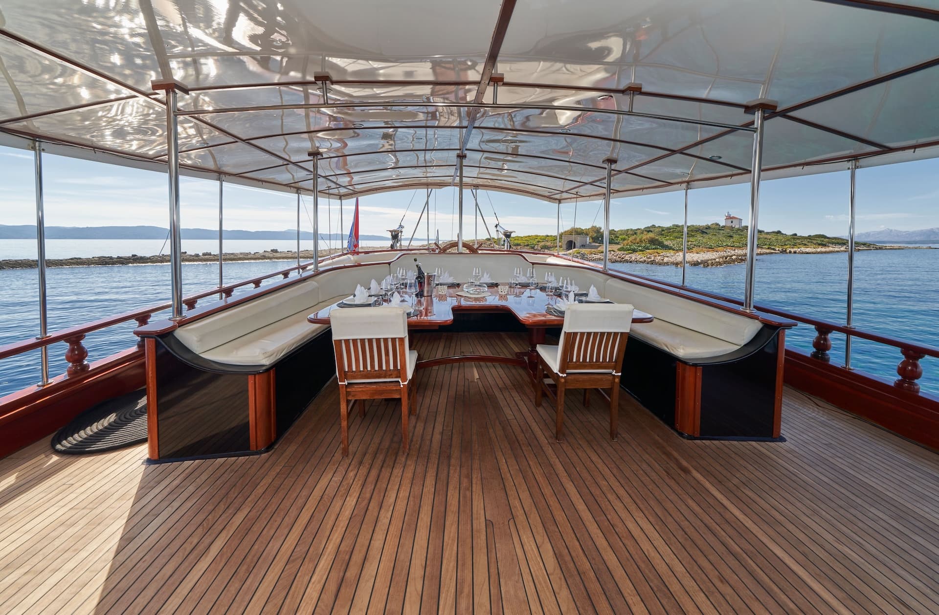 Dining area on wooden boat deck overlooking rocky island with small white lighthouse.