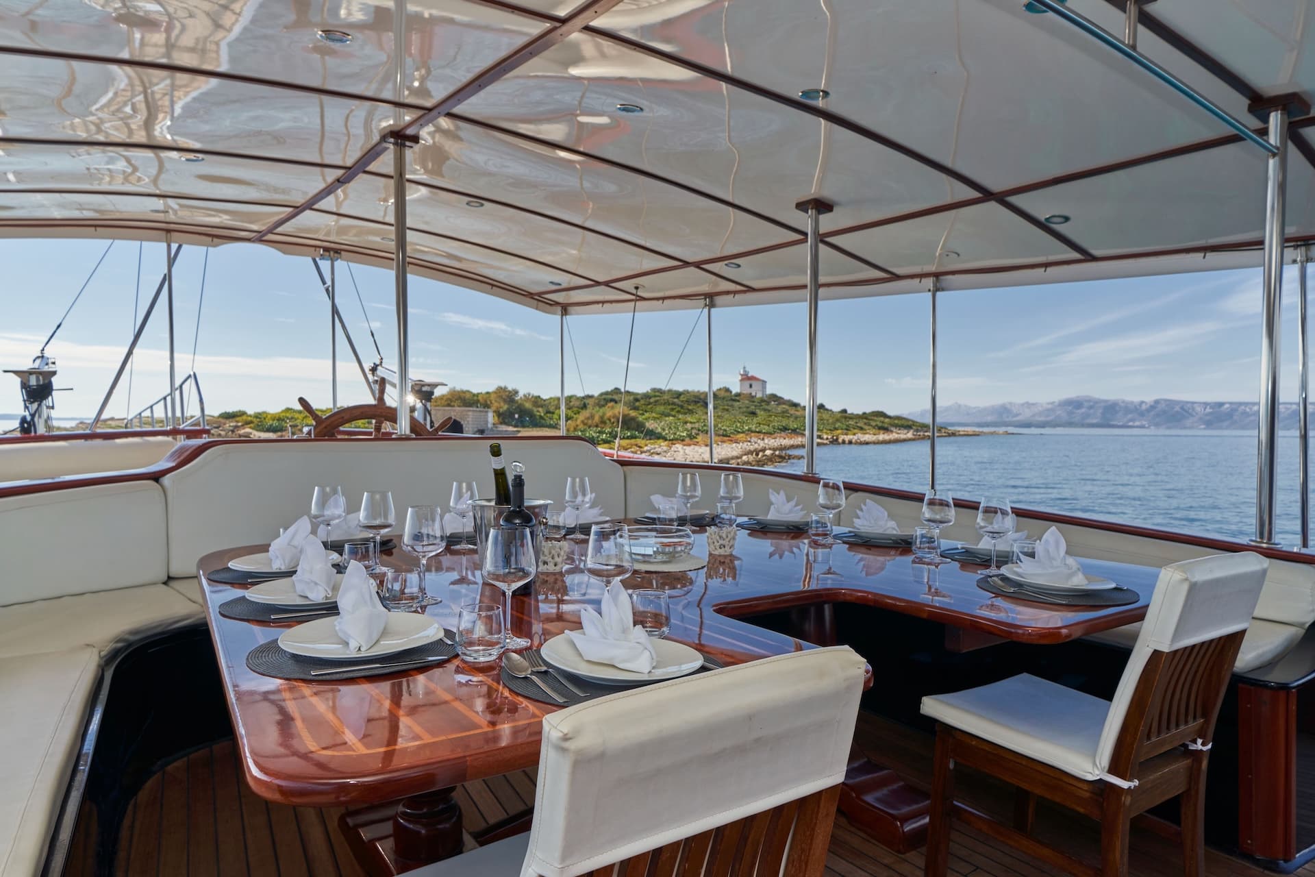 Outdoor dining area on yacht with set table overlooking island and distant mountains.