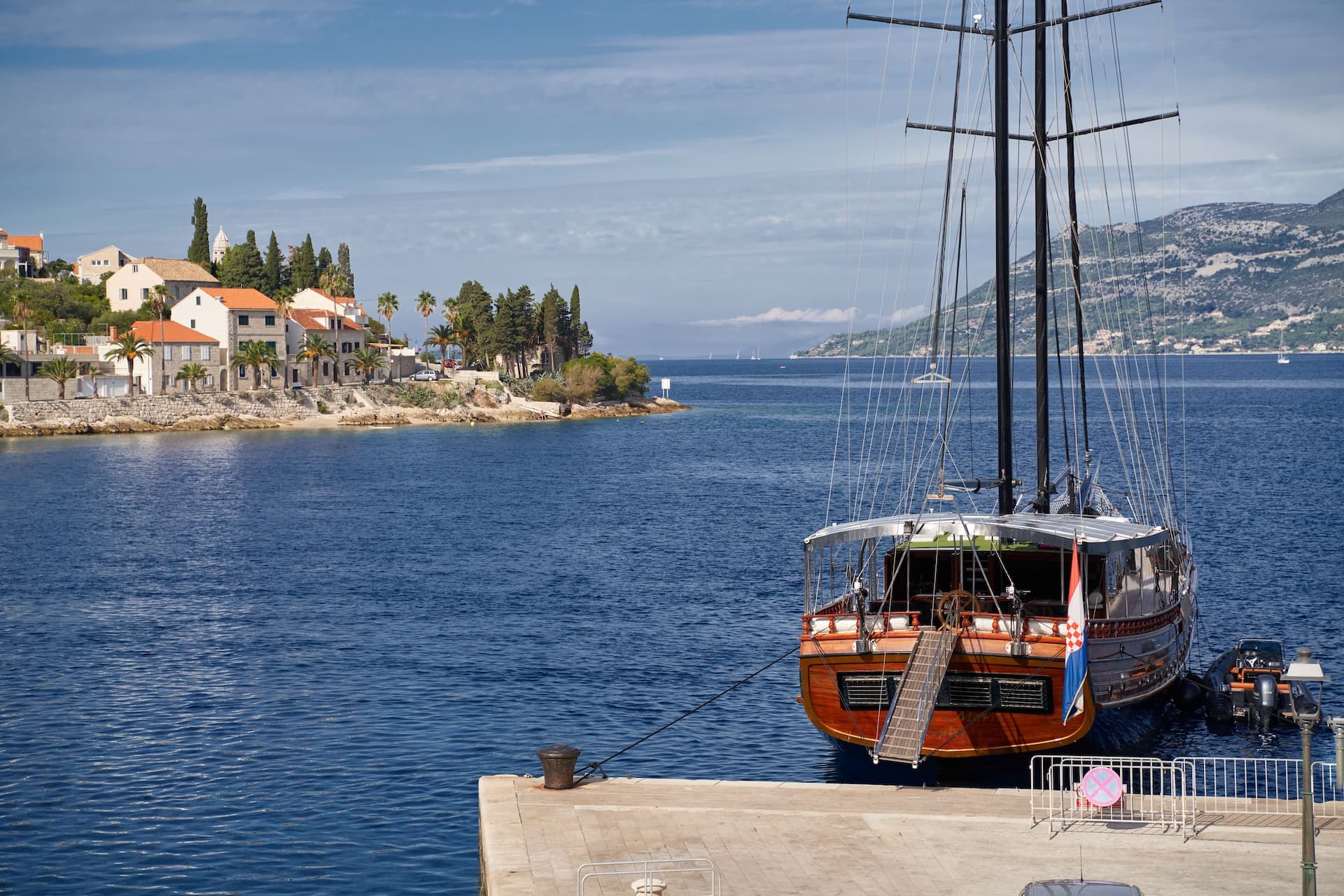 Wooden sailboat docked at pier with Croatian flag, coastal town, and mountains in background.