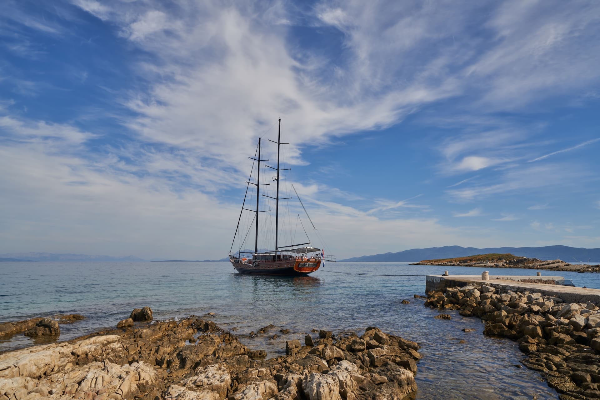 Large sailboat docked near rocky shore and small stone pier under blue sky with wispy clouds.