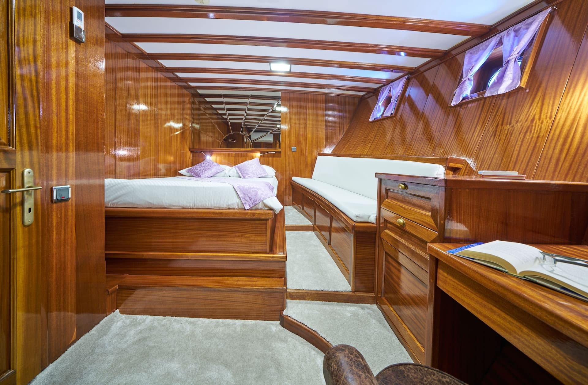 Cabin interior with glossy wood paneling, bed, white seating, and desk area.