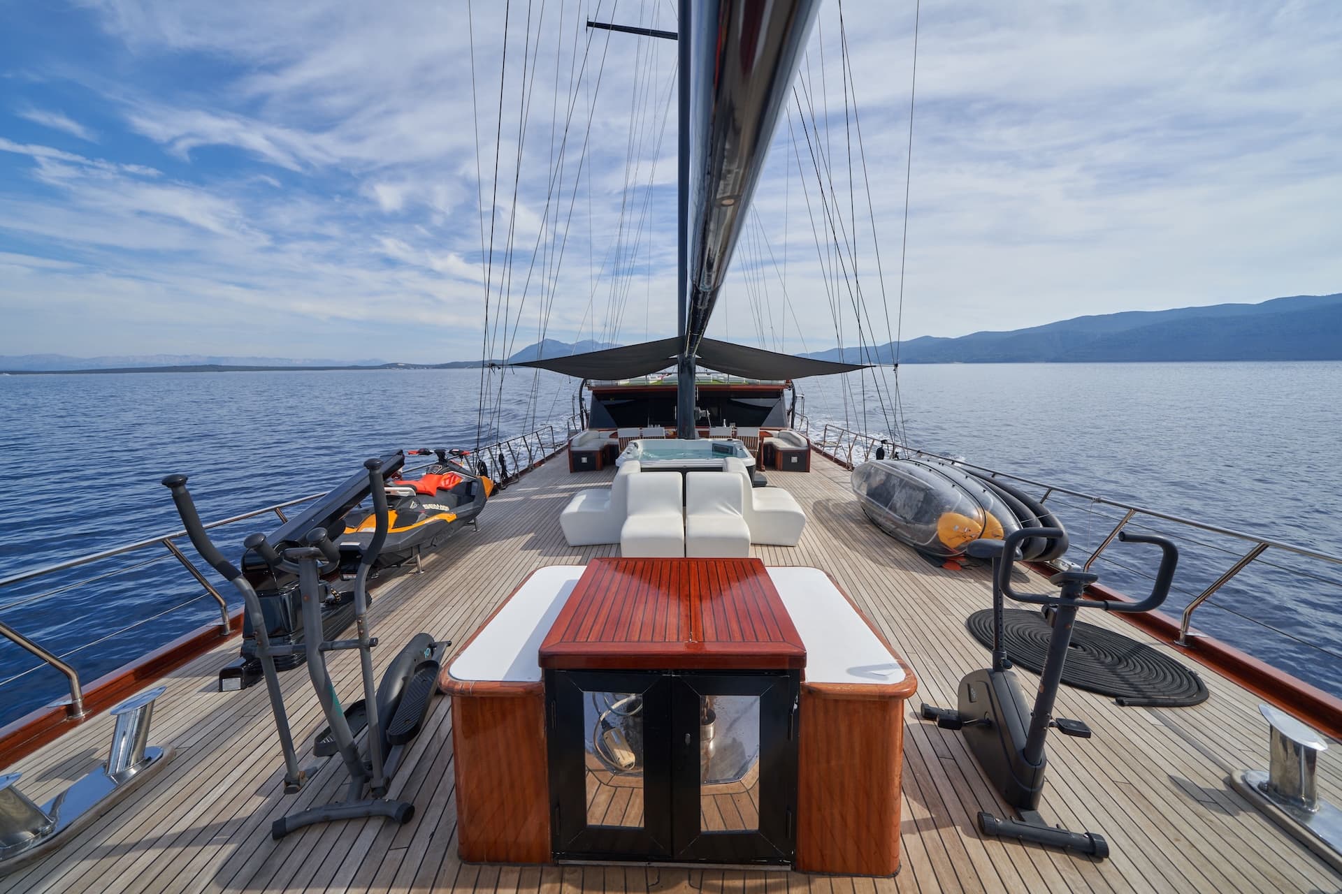 Yacht deck with exercise equipment, jet ski, and hot tub sailing near mountains.