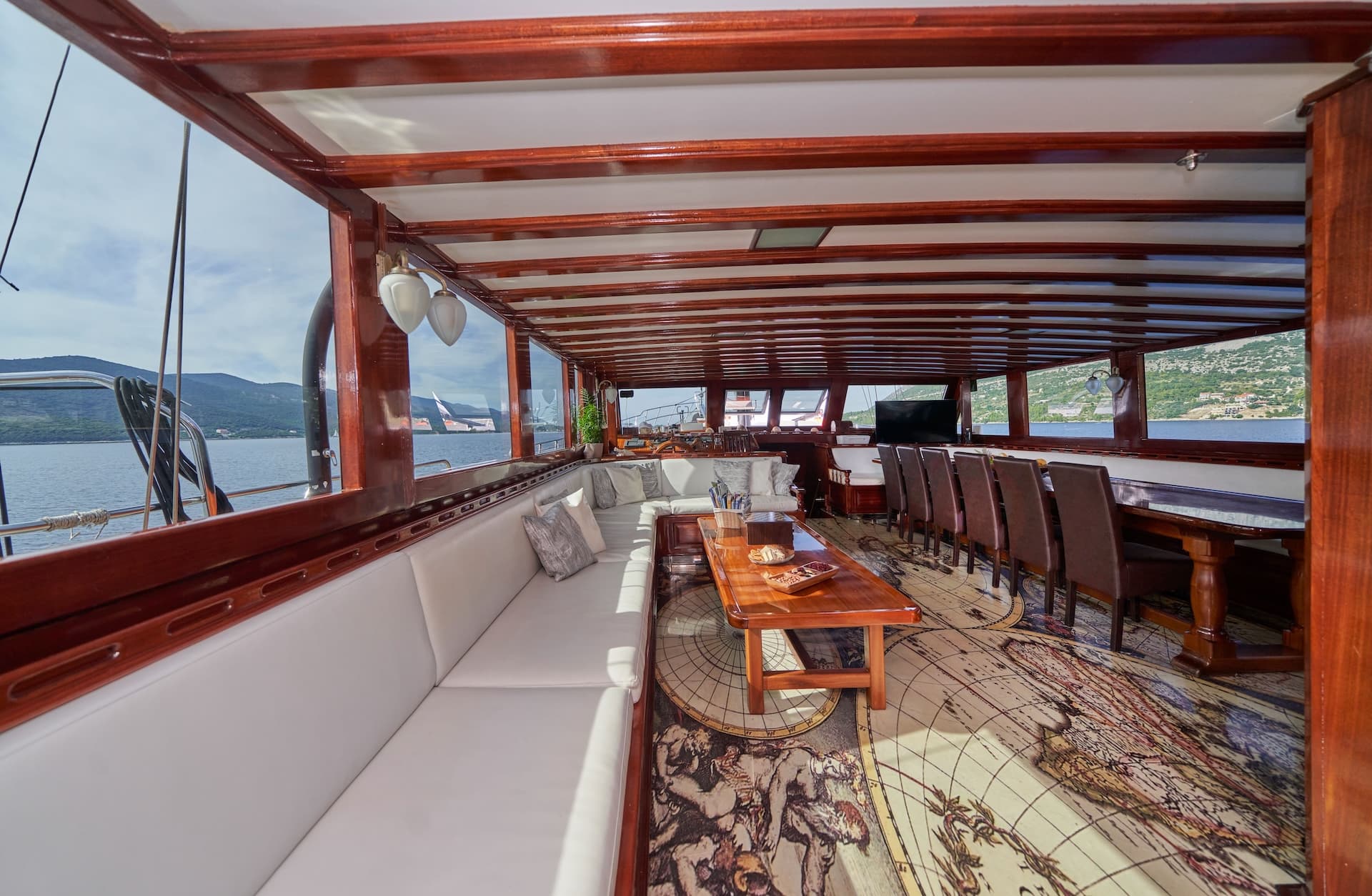 Indoor lounge area of a wooden boat with white seating and a map-themed rug, cruising past mountains.