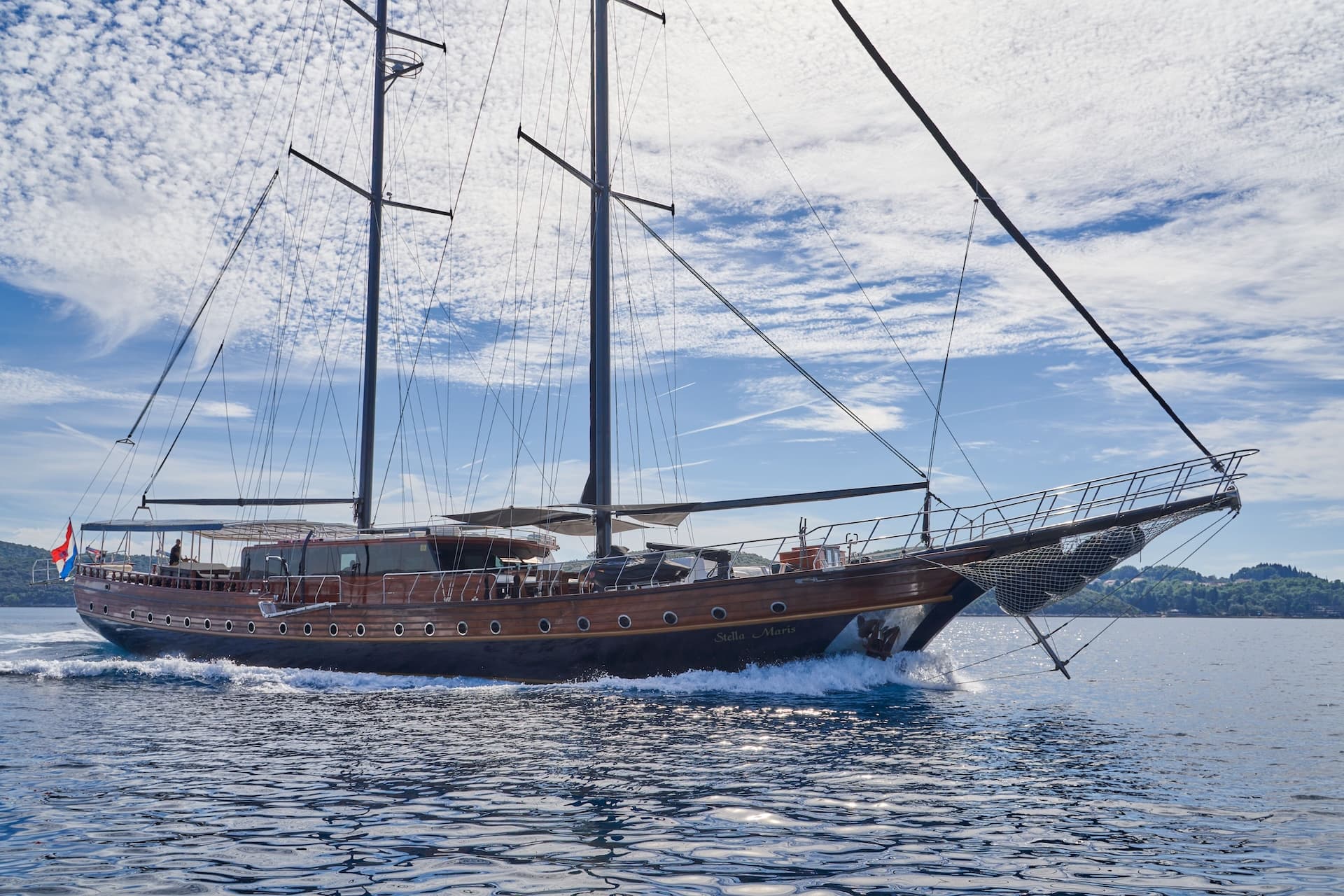 Wooden sailing yacht Stella Maris moving fast on blue water near a green coastline under a cloudy sky.