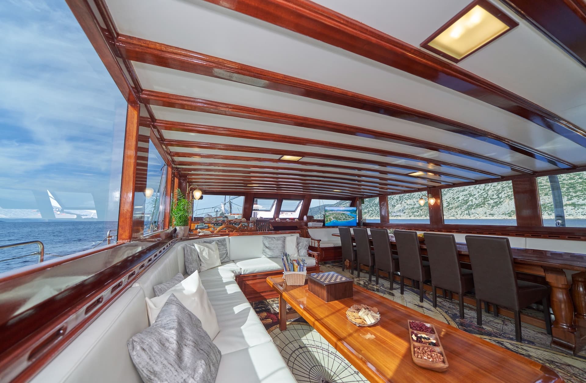 Salon area of a yacht with white seating, wood trim, and views of a rocky coastline.