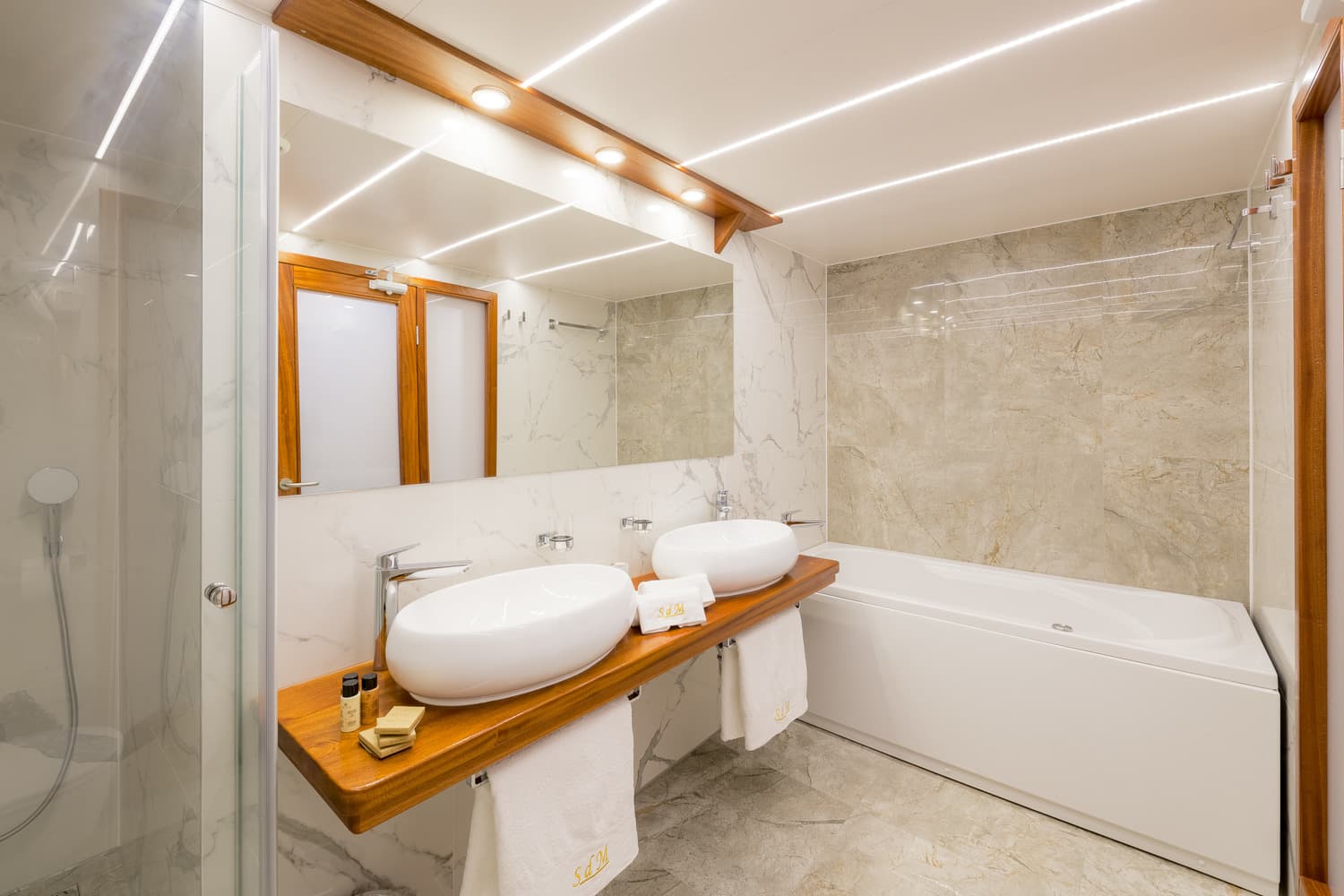 Modern bathroom with dual vessel sinks, wood vanity, and white bathtub with marble tile.