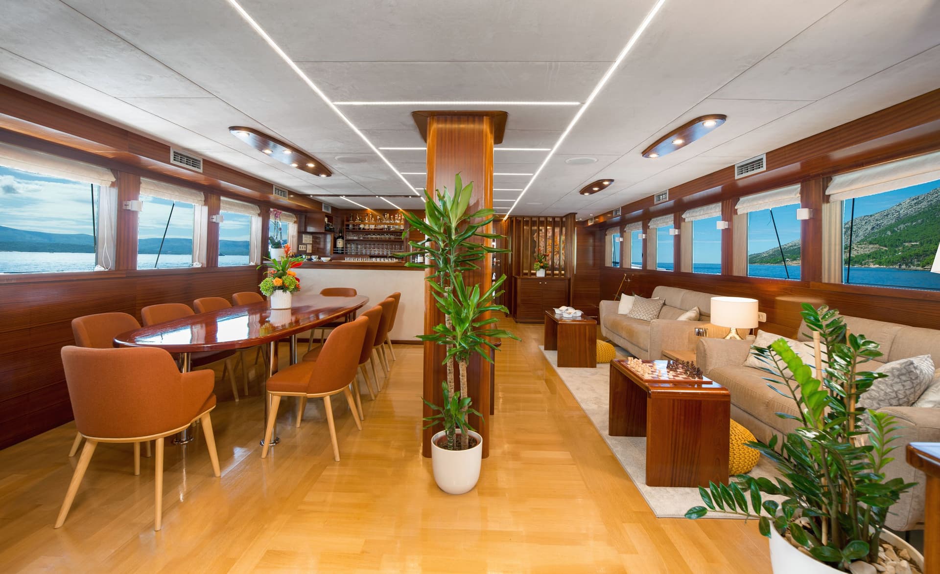 Interior lounge and dining area of a yacht with wood paneling and sea views of a coastline.
