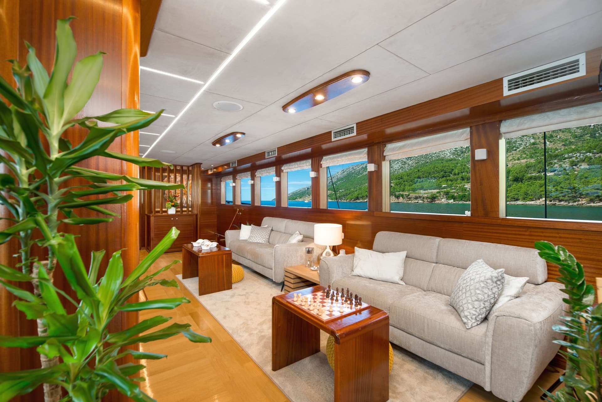 Lounge room interior with wood paneling, beige sofas, and sea view of green hills.