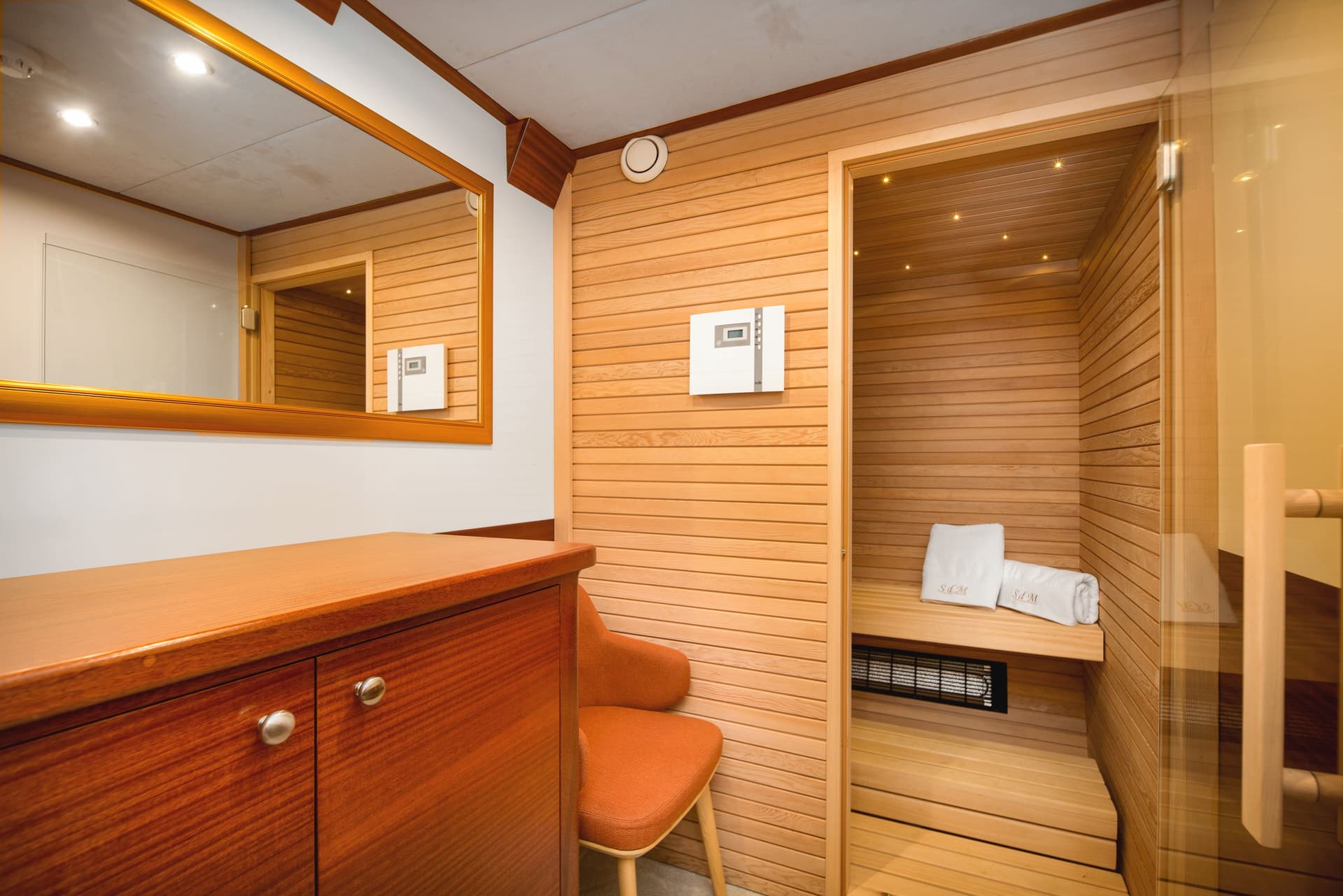 Interior view of a wooden sauna room with a glass door, next to a wooden cabinet and orange chair.