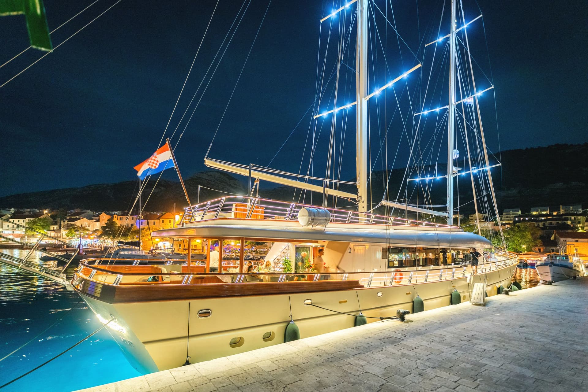 Large illuminated sailboat docked at night with Croatian flag near illuminated coastal town.