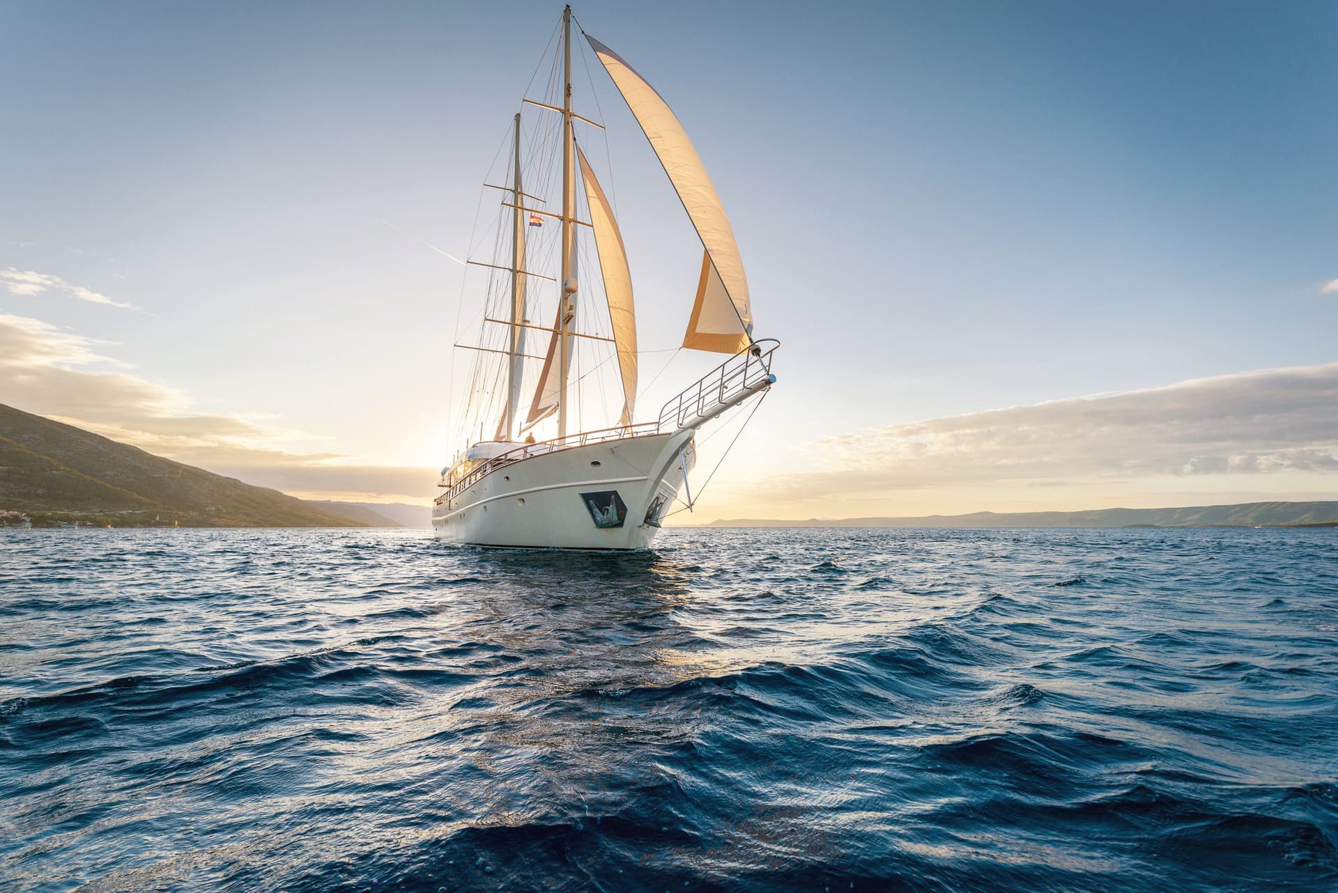 Large sailboat with sails open on deep blue water near a hilly coastline at sunset.
