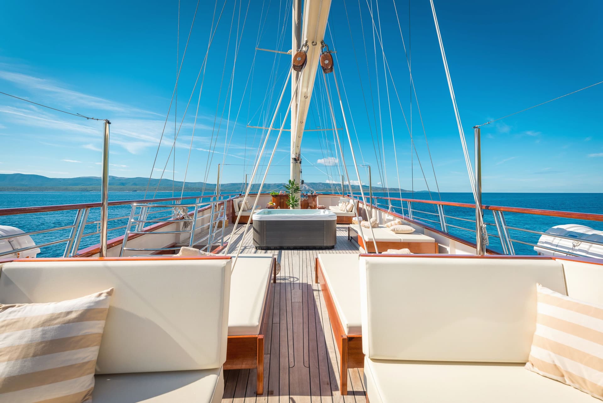 Yacht deck with hot tub, lounge seating, and clear blue water with distant mountains.
