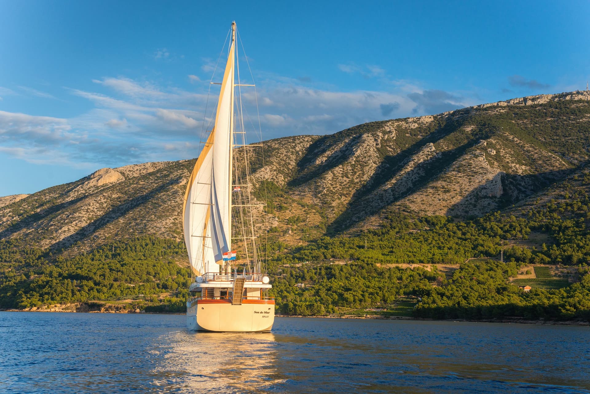Sailboat "Son de Mar" sailing near a steep, scrub-covered coastline in Croatia.
