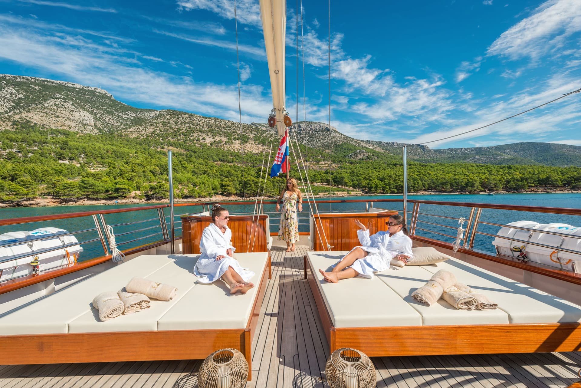 Women relaxing on sunbeds aboard a yacht near a lush, green, rocky coastline under a blue sky.
