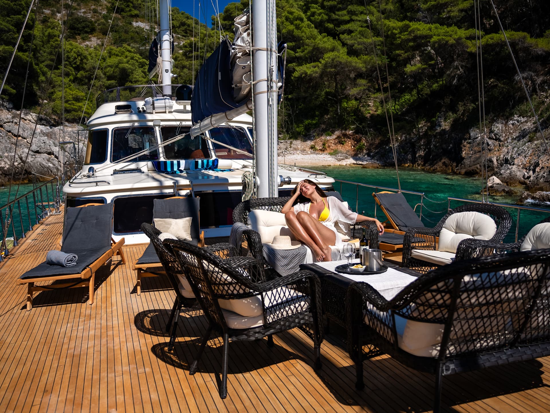 Woman relaxing on yacht deck near tree-covered rocky coastline with turquoise water