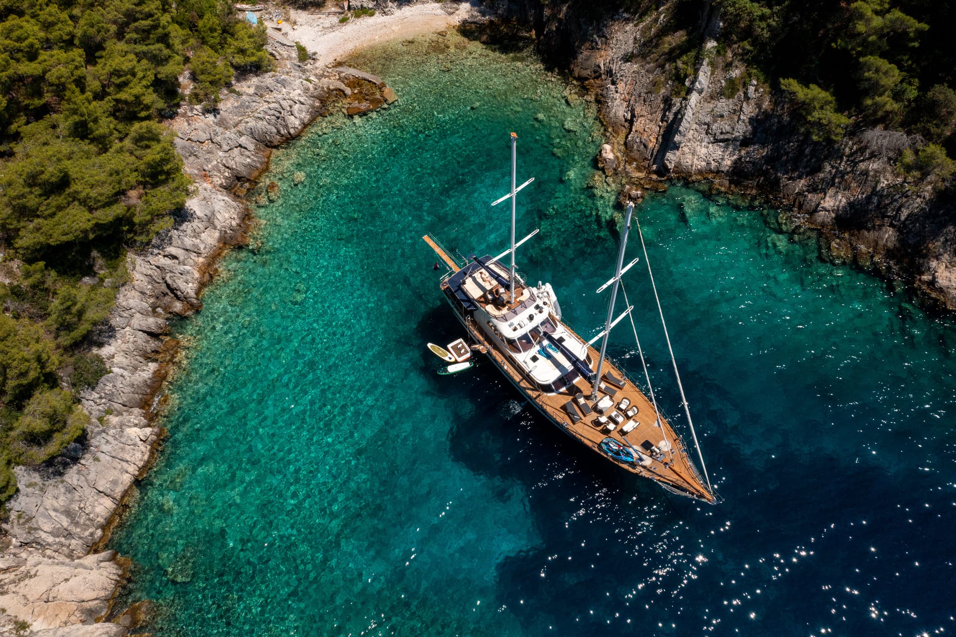 Sailing yacht anchored in turquoise cove near rocky, tree-covered coastline.