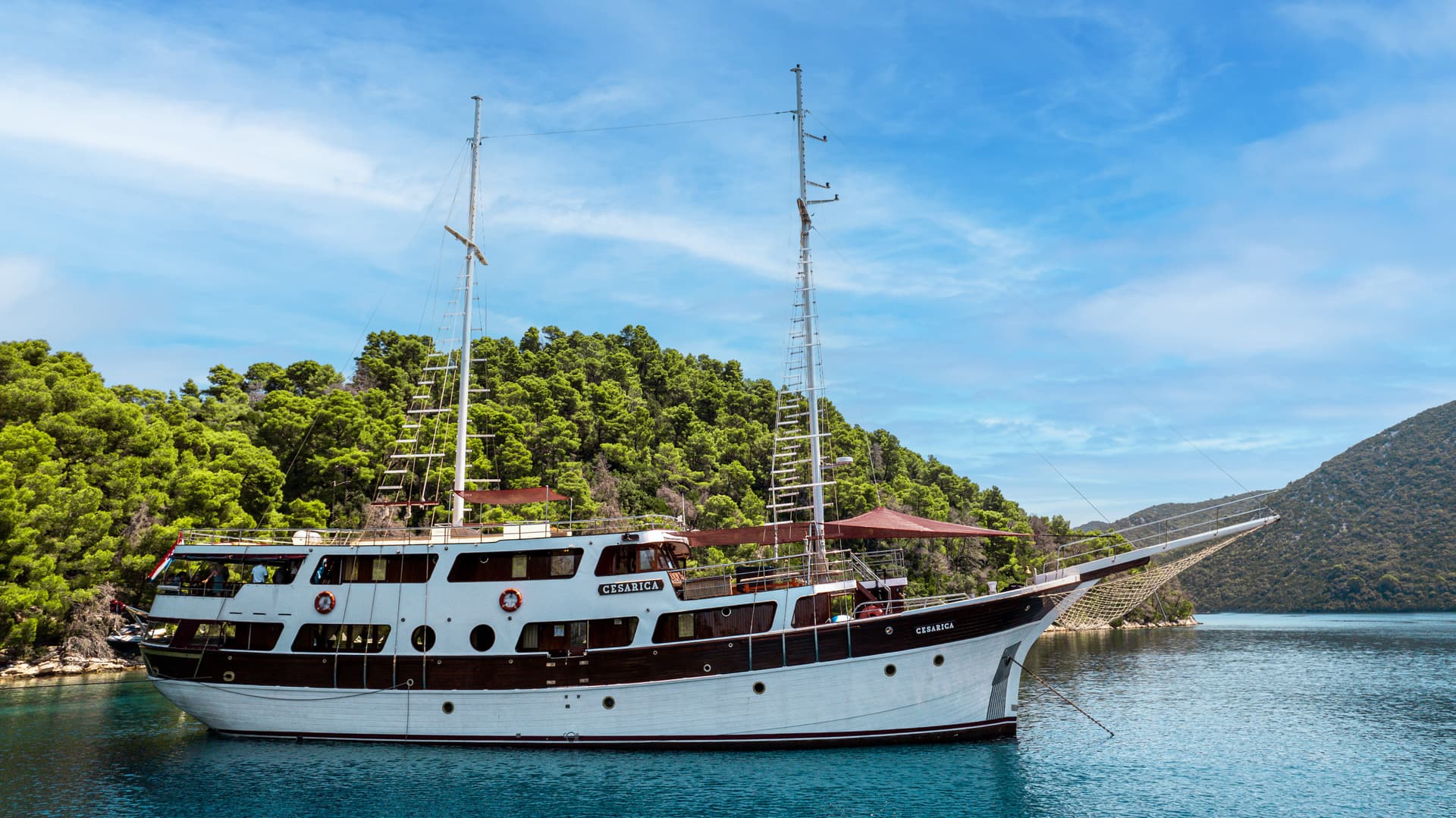 Motor sailor boat 'Cesarica' anchored in blue water near a densely forested coastline under a blue sky.