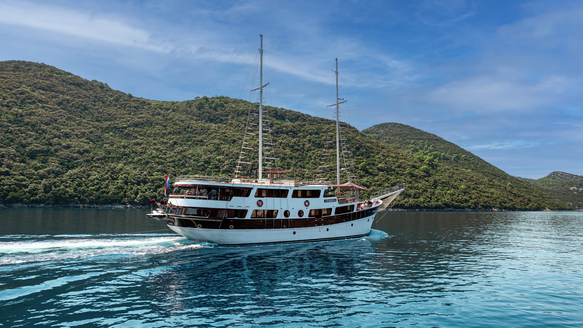 White and brown two-masted boat named Cesarica cruising on blue water near forested hills.
