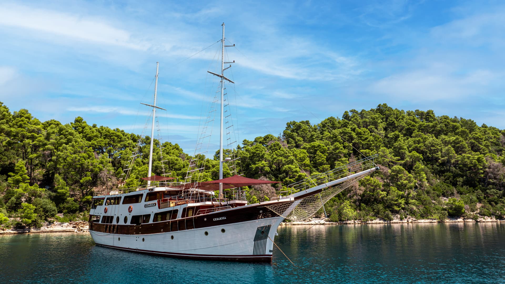 Motor sailer "Cesarica" anchored in calm blue water near a densely forested coastline.