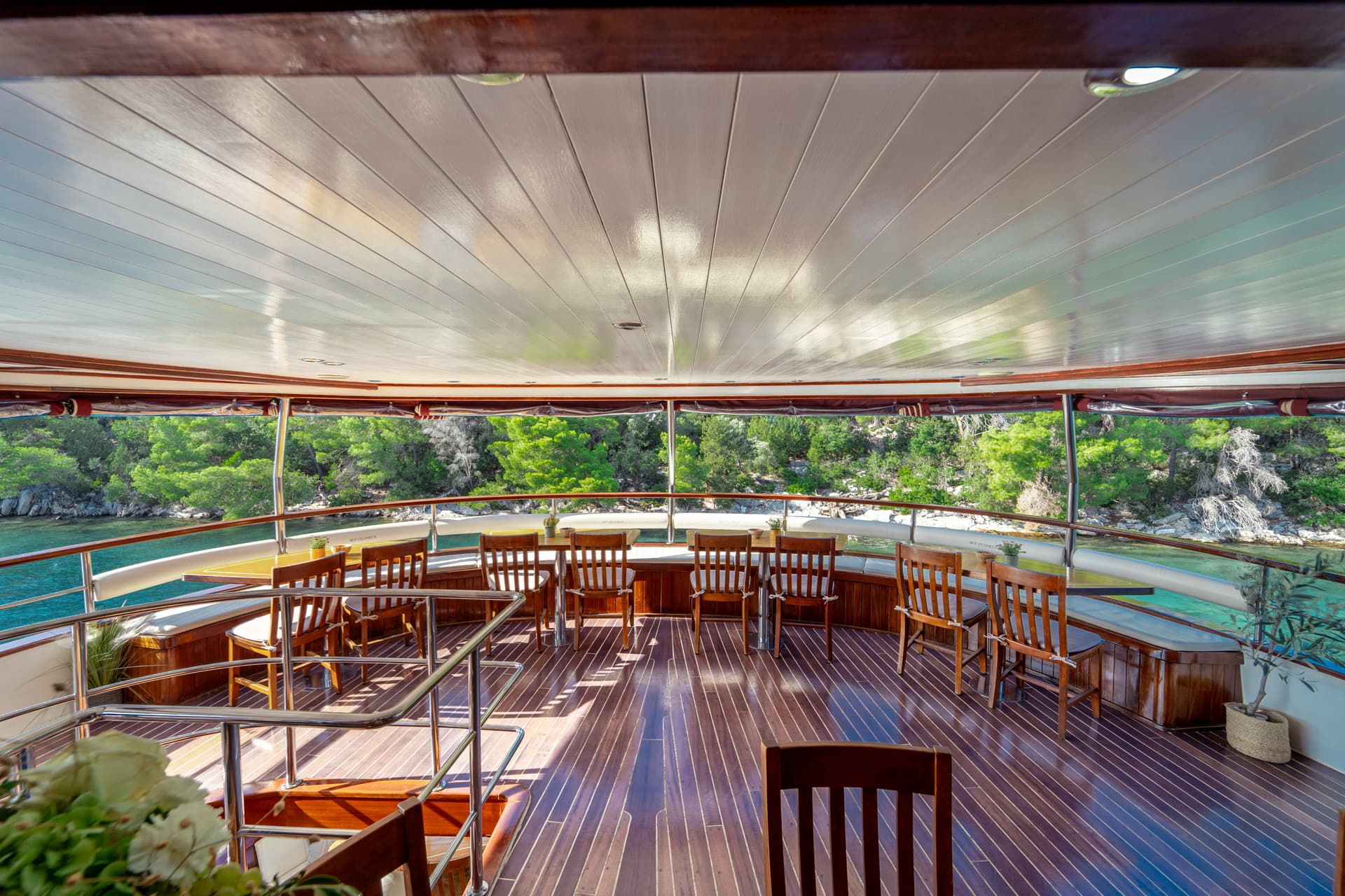 Covered dining area on a wooden boat deck overlooking turquoise water and green coastline.
