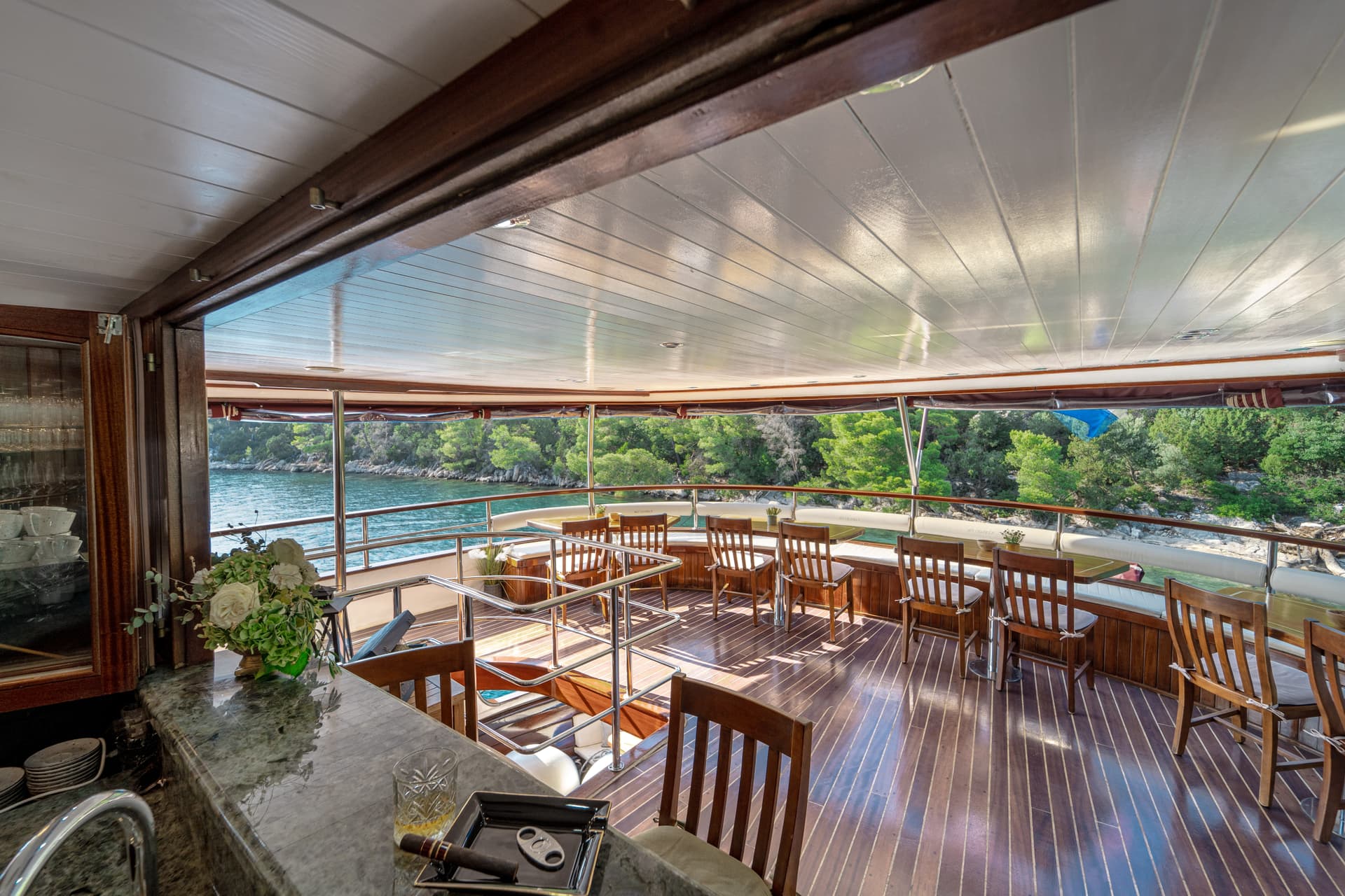 Boat deck bar area with wooden chairs overlooking a lush, tree-lined coastline and blue water.