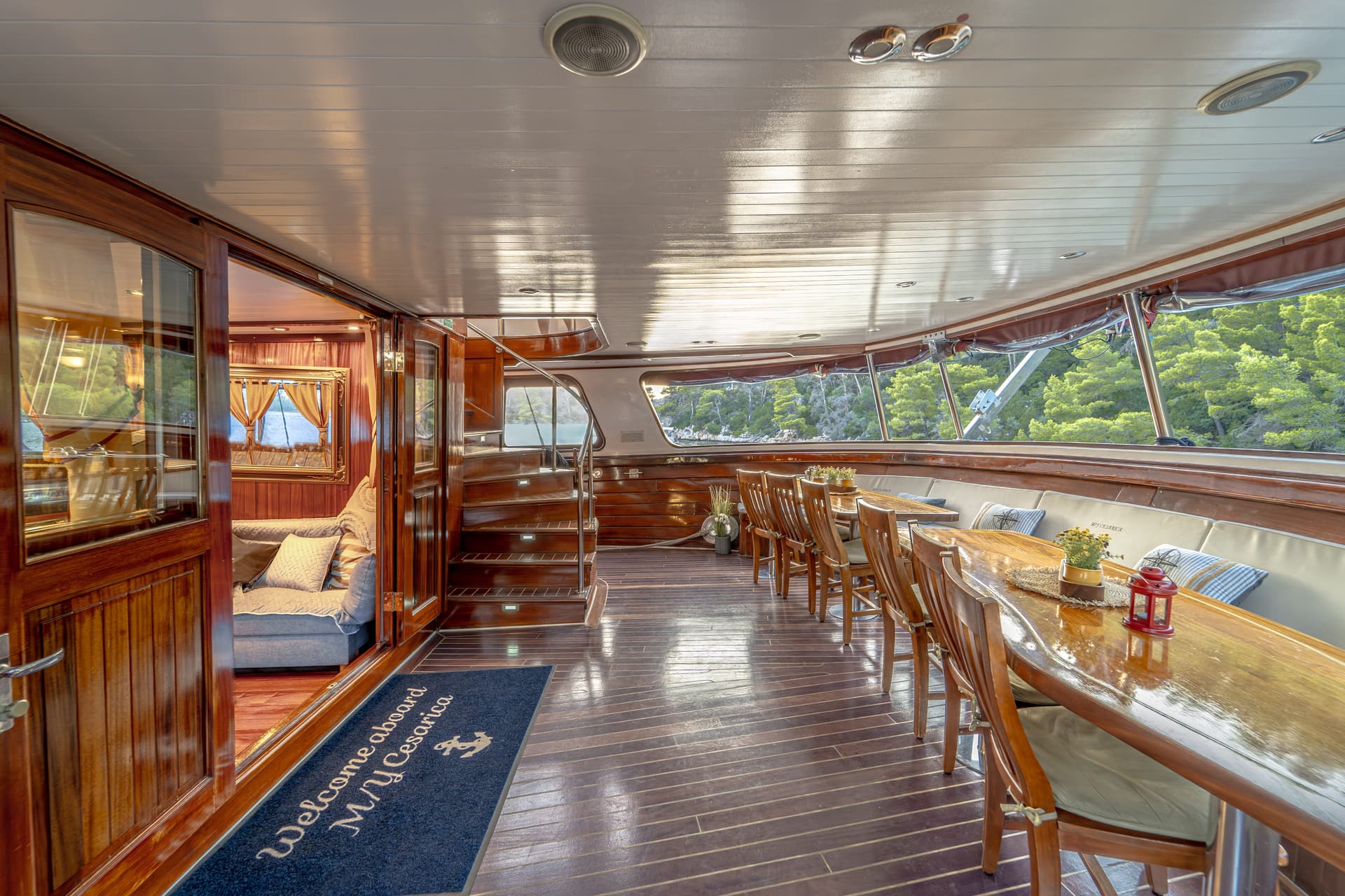 Interior dining area of M/Y Cesárica yacht with wood paneling and view of forested coastline.