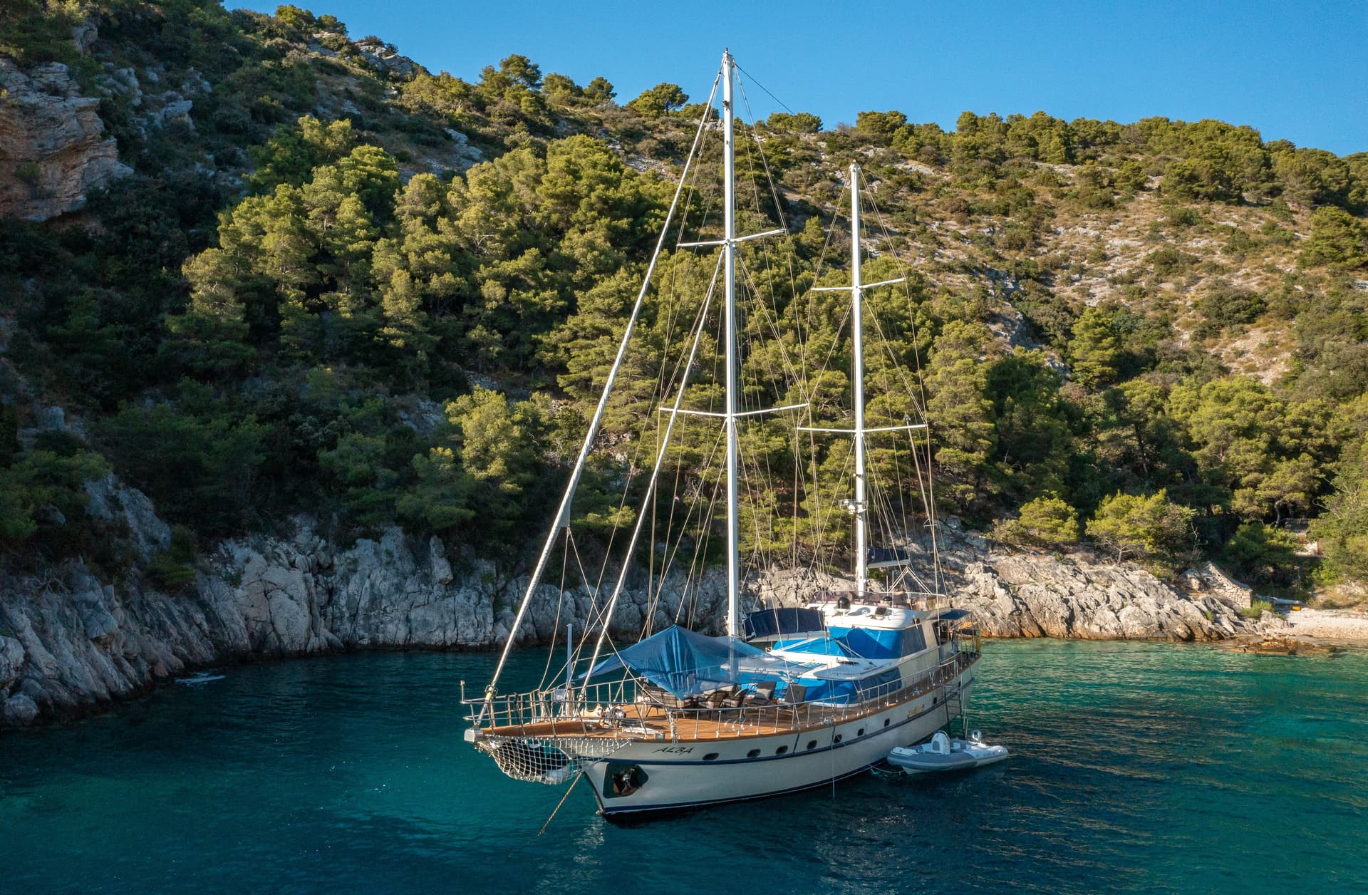 Large sailboat anchored in turquoise cove near rocky, forested coastline under clear blue sky