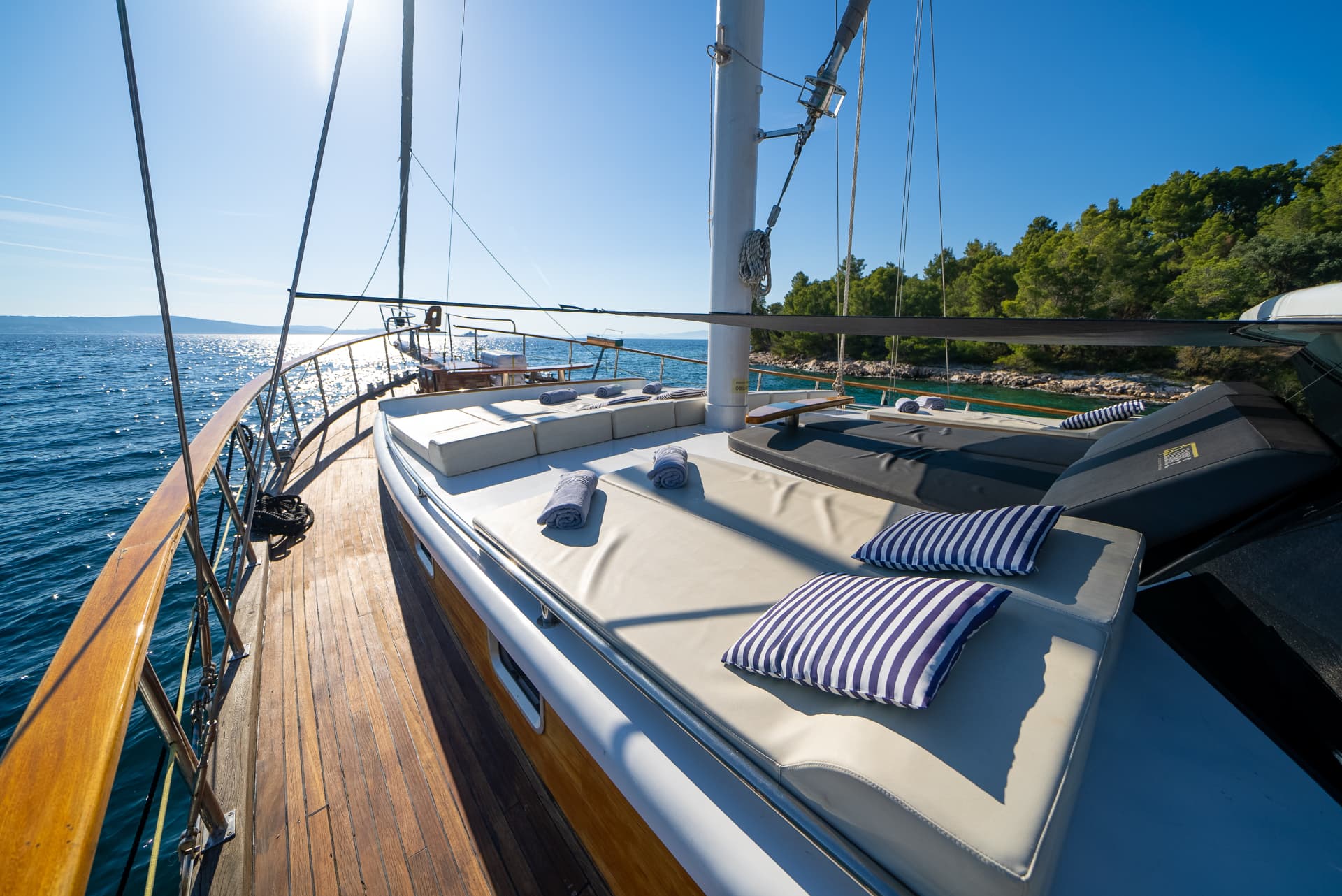 Deck of yacht with sunbeds and striped pillows near forested coastline on sunny day
