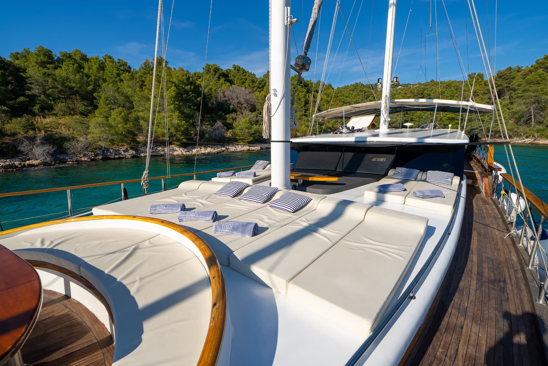 Yacht sun deck with white cushions and striped pillows moored near forested coastline