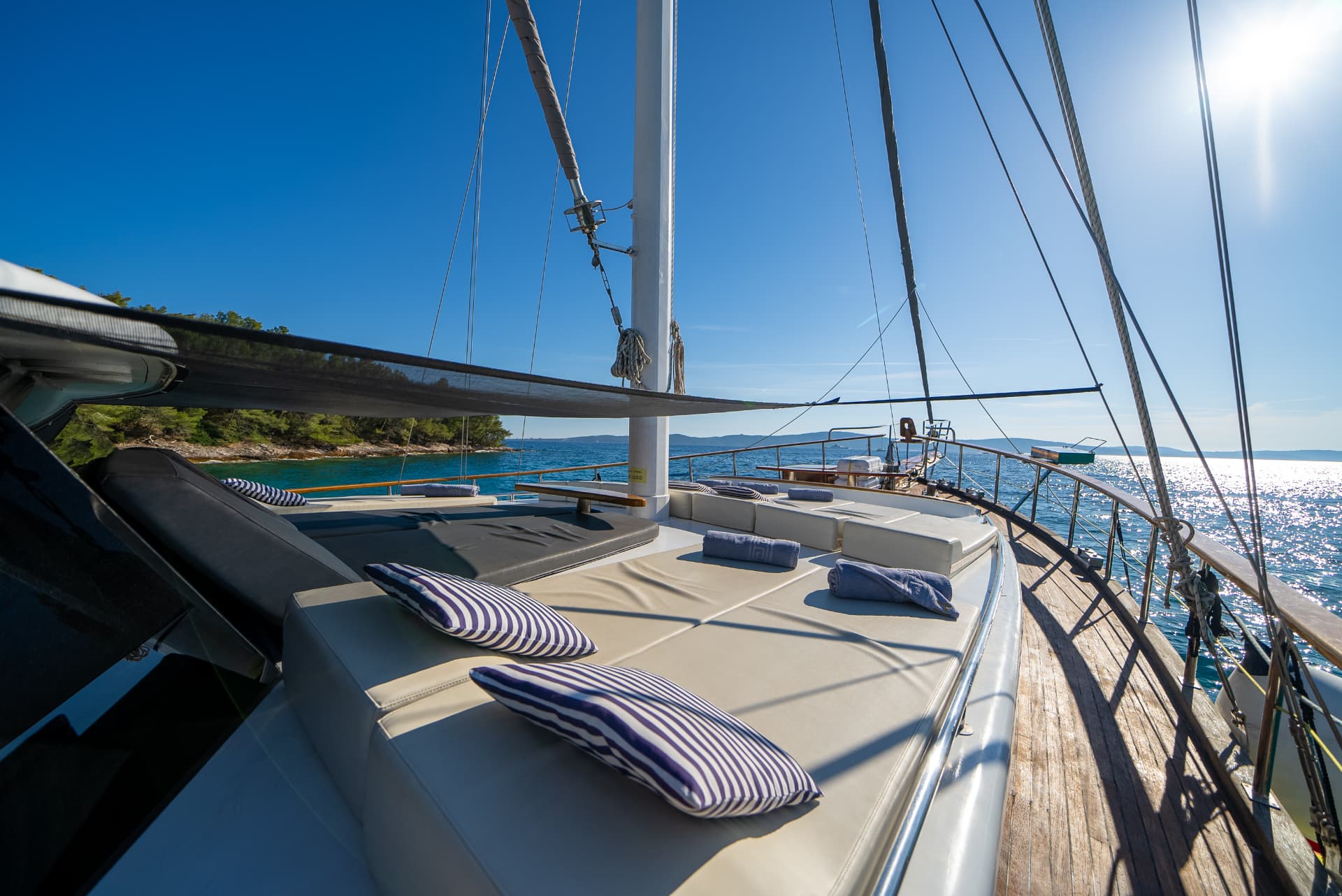 Sun deck with striped pillows on yacht near forested coastline under bright blue sky.