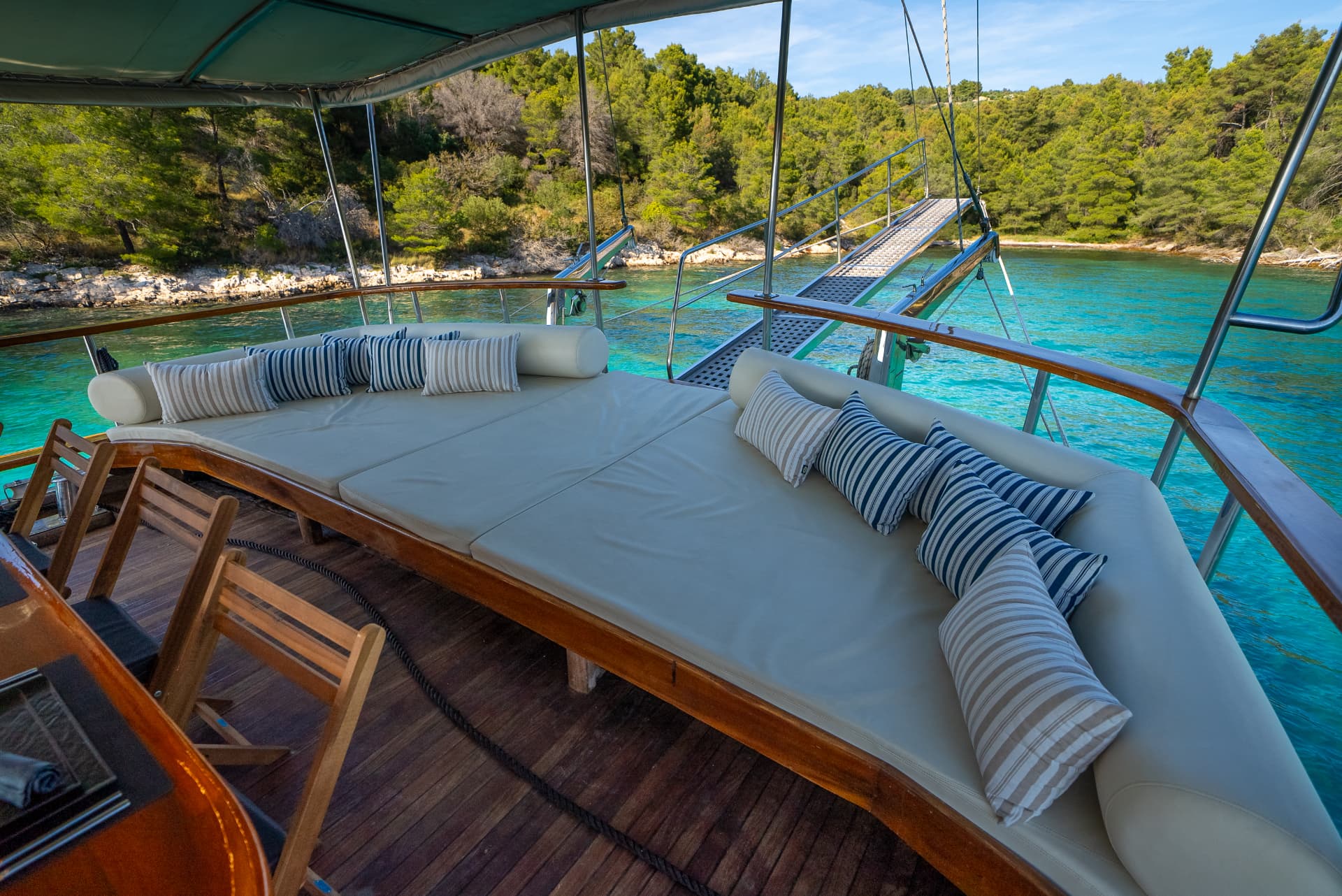 Boat deck with sunbed and striped pillows anchored near forested coastline with turquoise water.