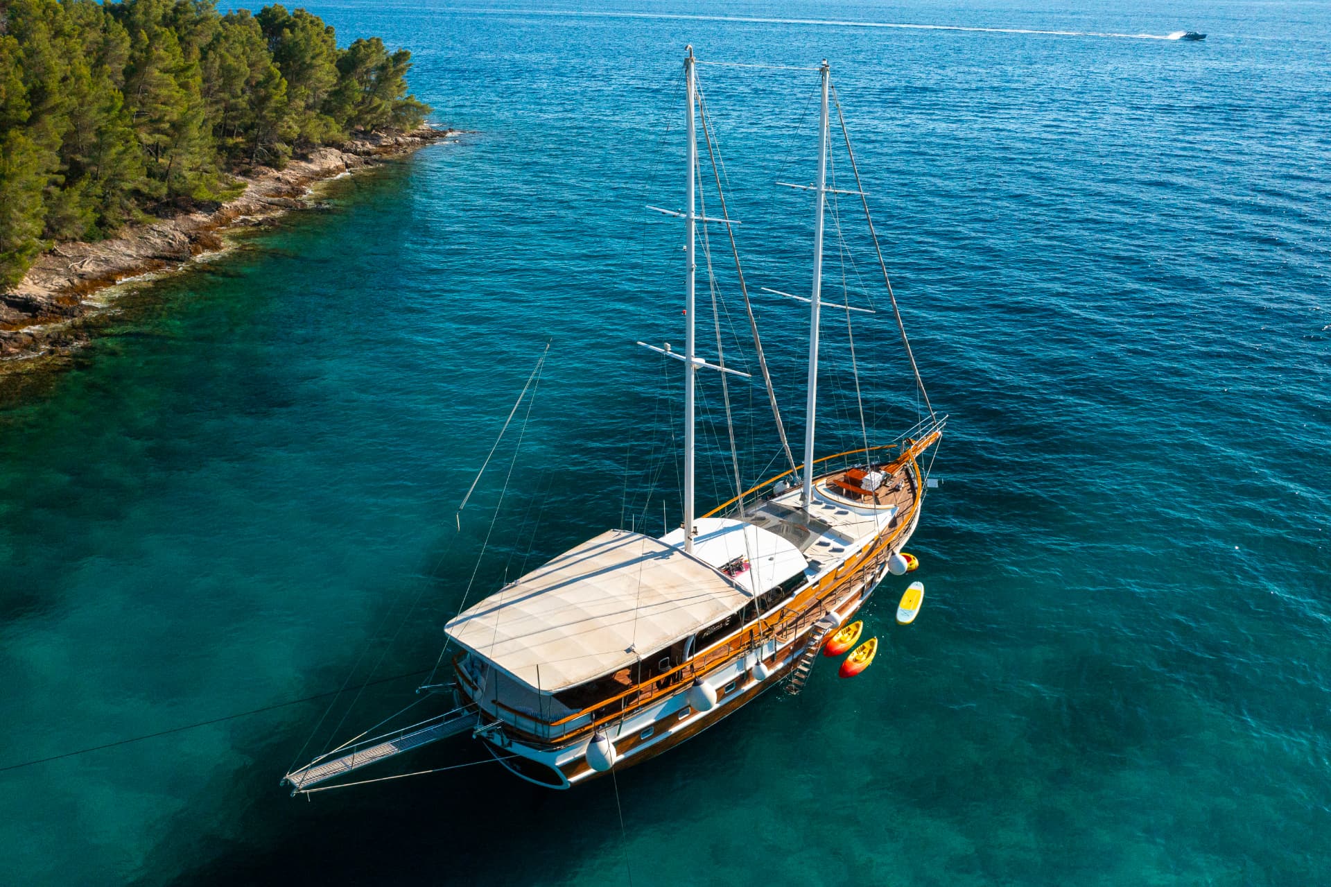 Large wooden sailboat anchored near a rocky, tree-lined coast with clear blue water