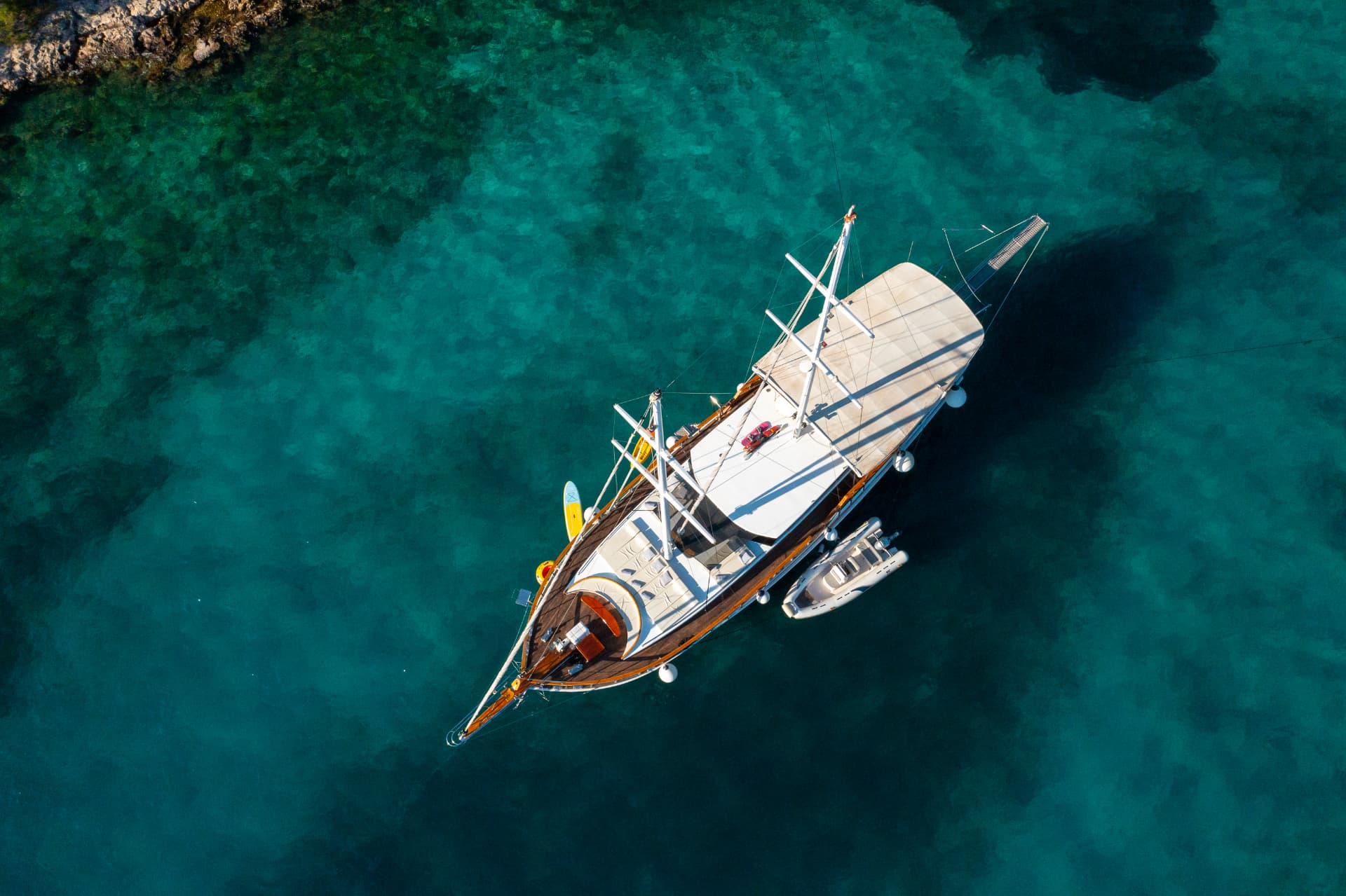 Large sailboat anchored near rocky coast in clear turquoise water, aerial view