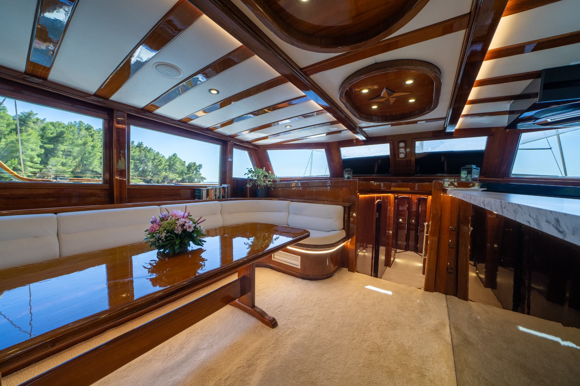 Yacht interior with glossy wood, white seating, and view of green trees from windows