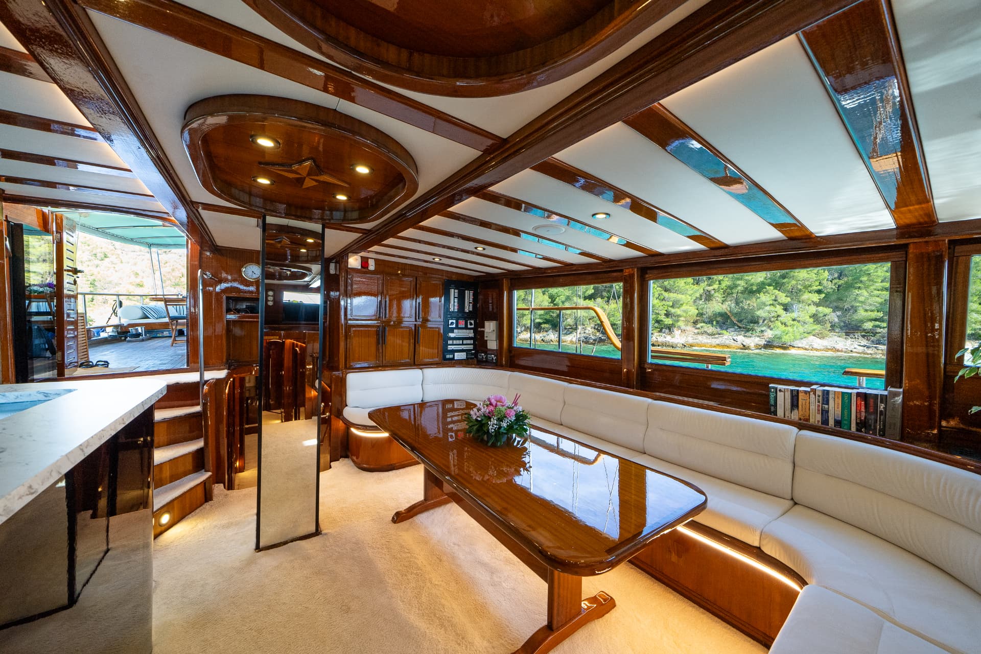 Yacht interior with glossy wood, white seating, and view of forested coastline on turquoise water.