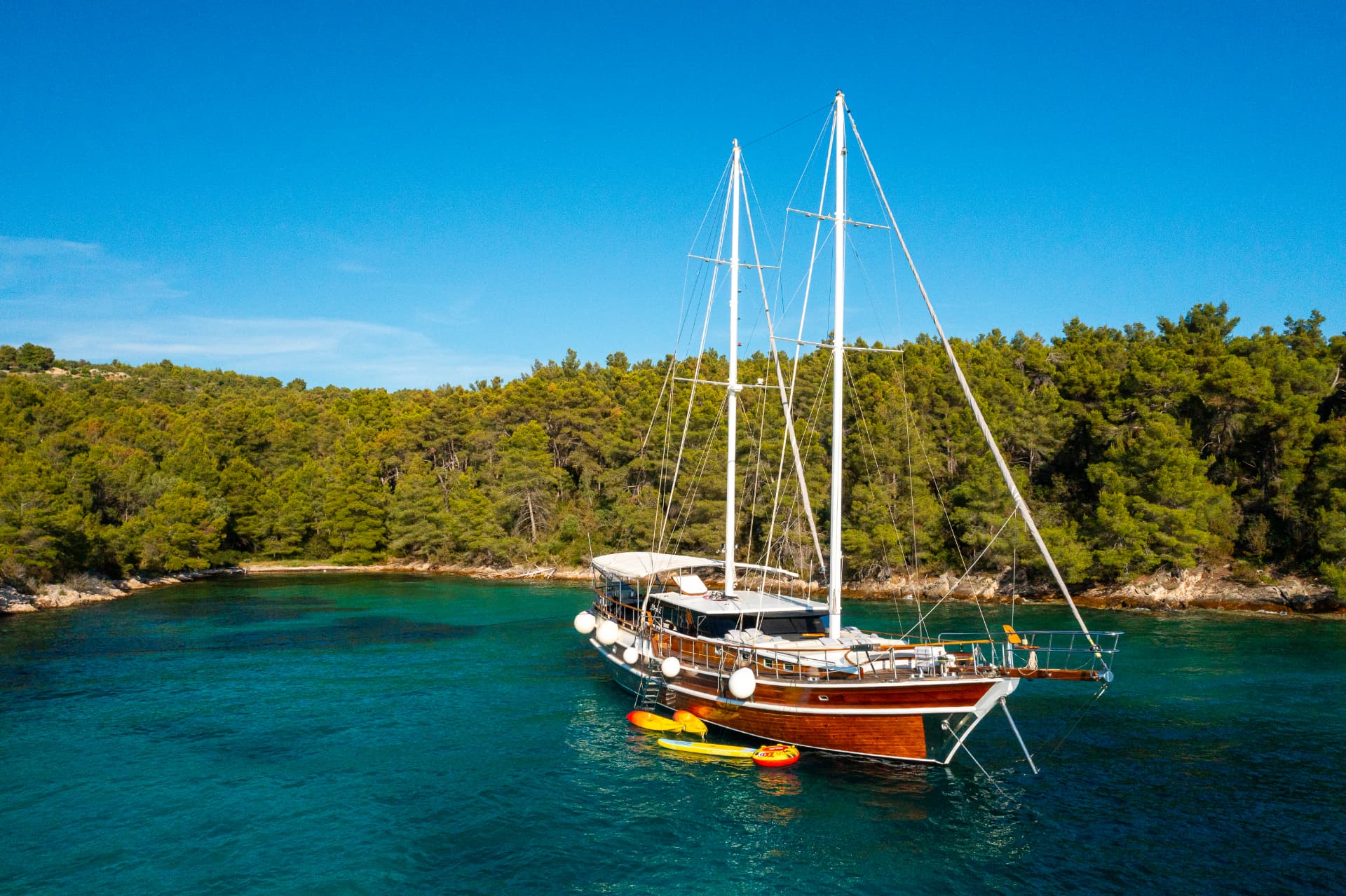 Wooden sailboat anchored in turquoise cove by dense green forested coastline