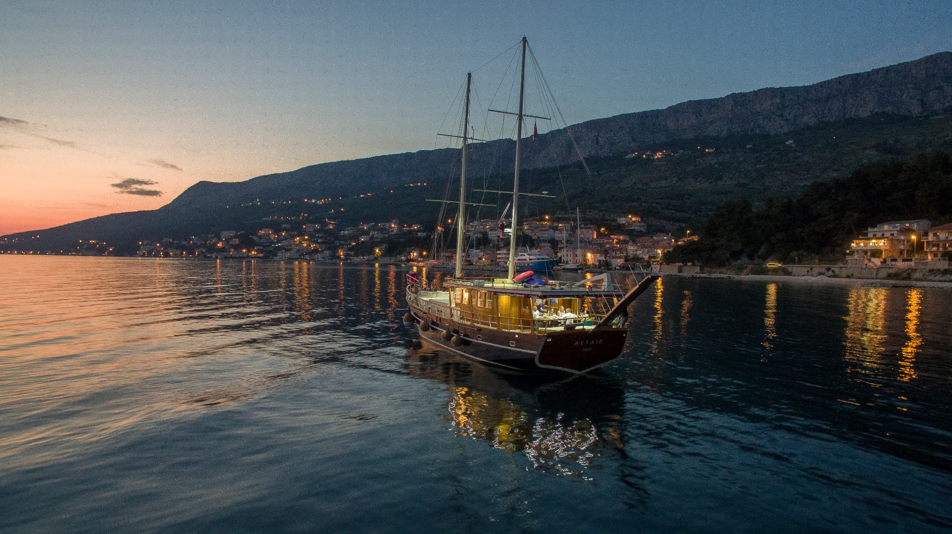 Illuminated sailboat on calm water near a coastal town at dusk with mountain backdrop