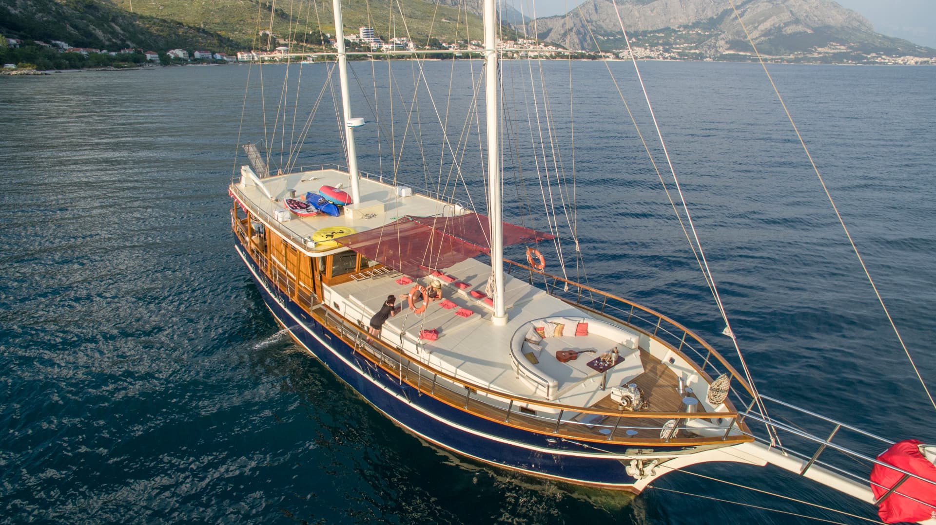 Large wooden sailboat on deep blue water near a mountainous coastline with buildings.