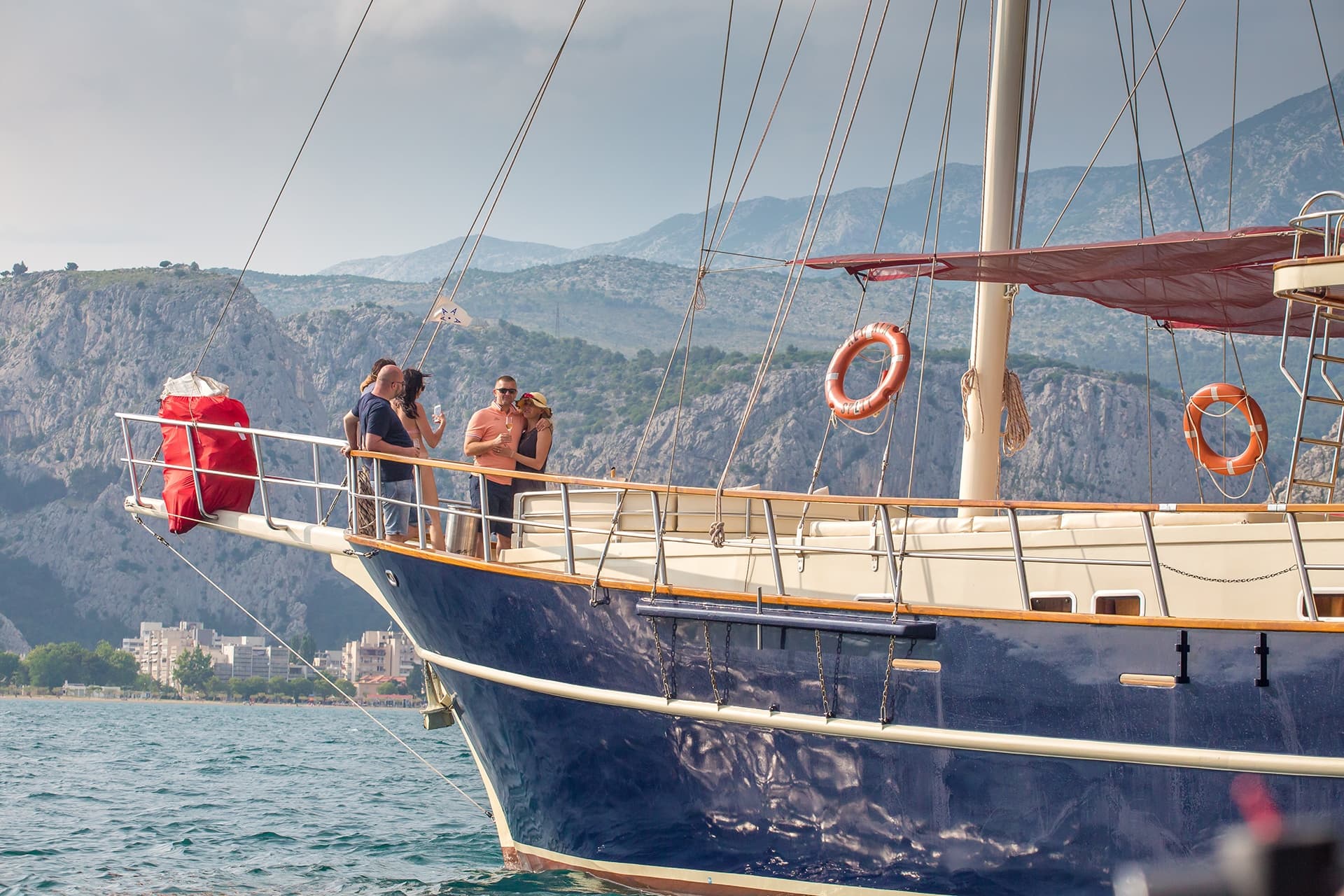 People on bow of dark blue sailboat with mountains and coastal town in background