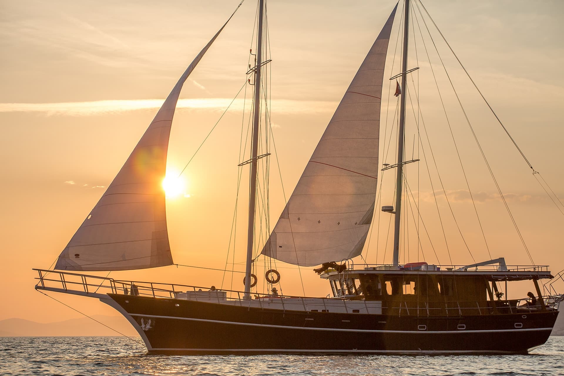 Large sailboat with sails up on the water during a bright orange sunset.