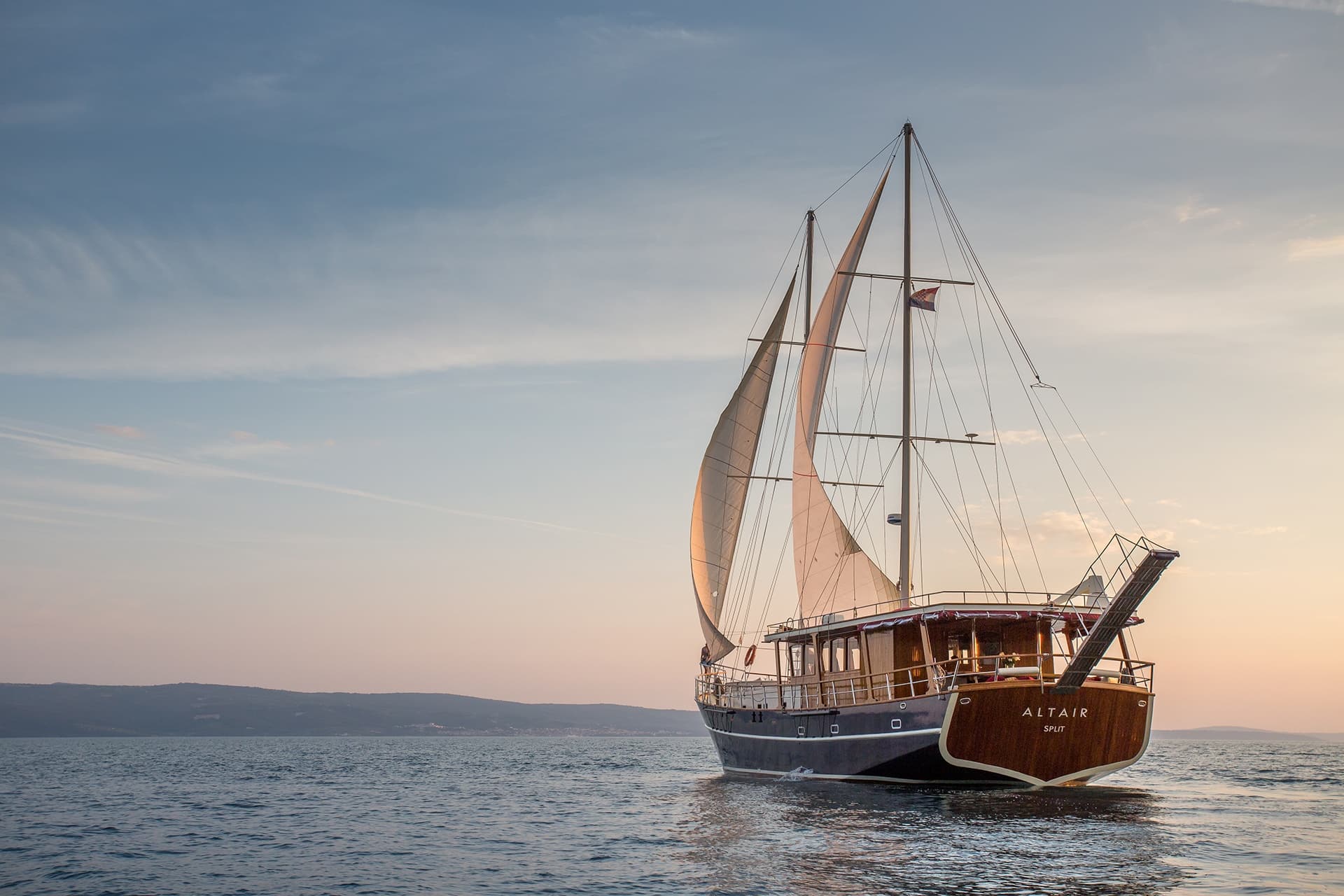Motor sailer Altair departing Split on calm sea at sunset with distant coastline