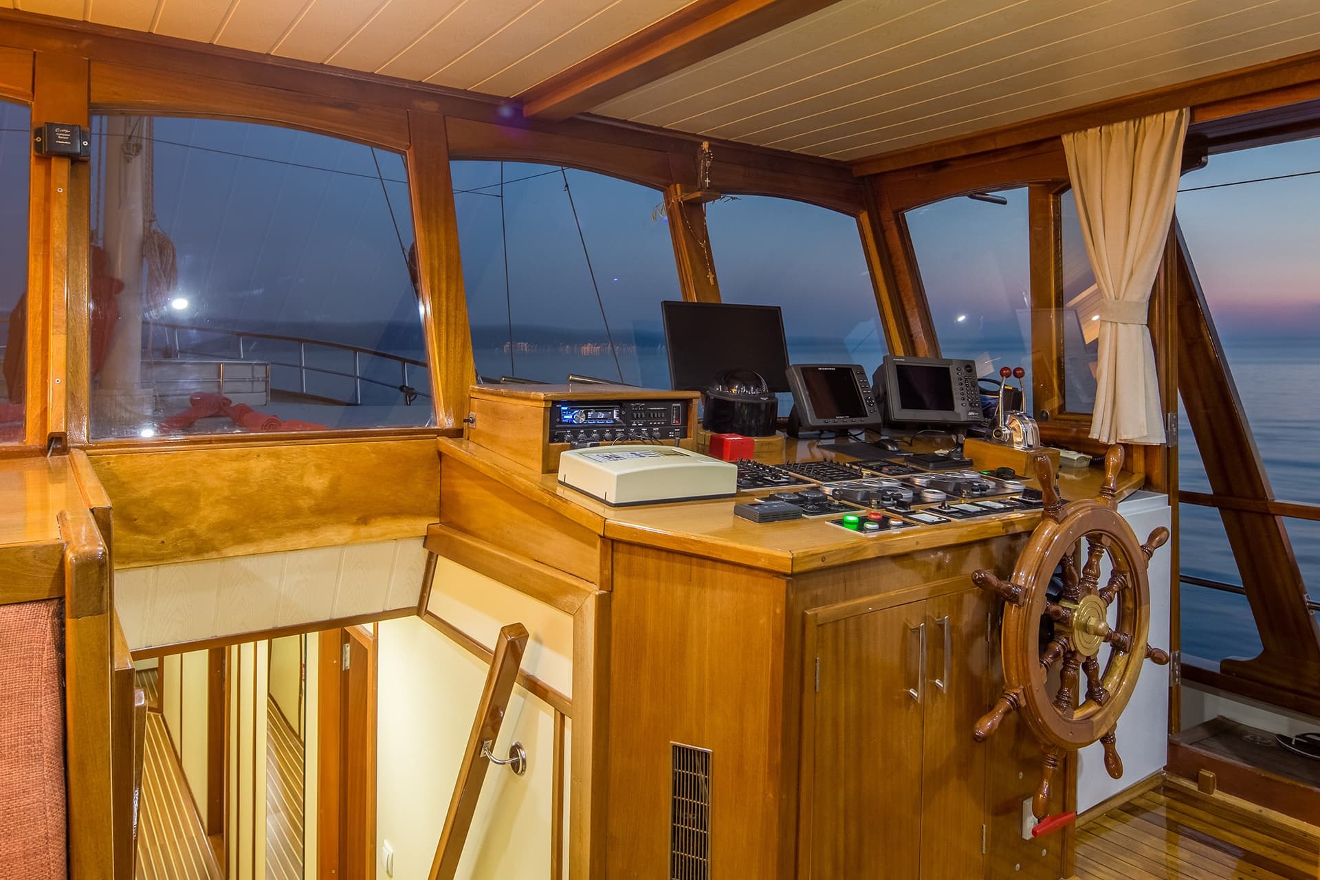 Yacht bridge interior with wooden steering wheel, navigation screens, and view of the sea at dusk.