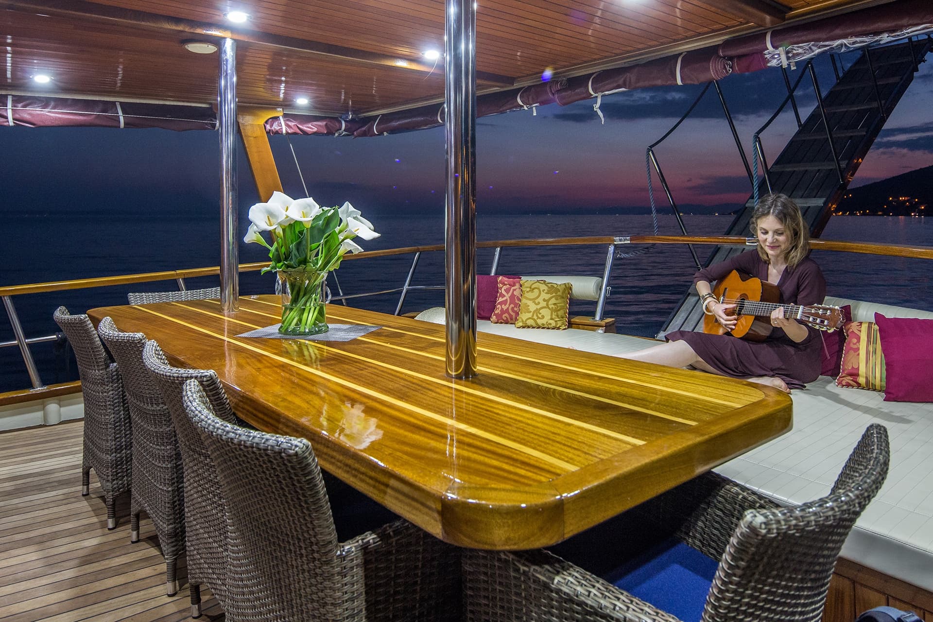Woman playing guitar on yacht deck with glossy wooden table and calla lilies at dusk.