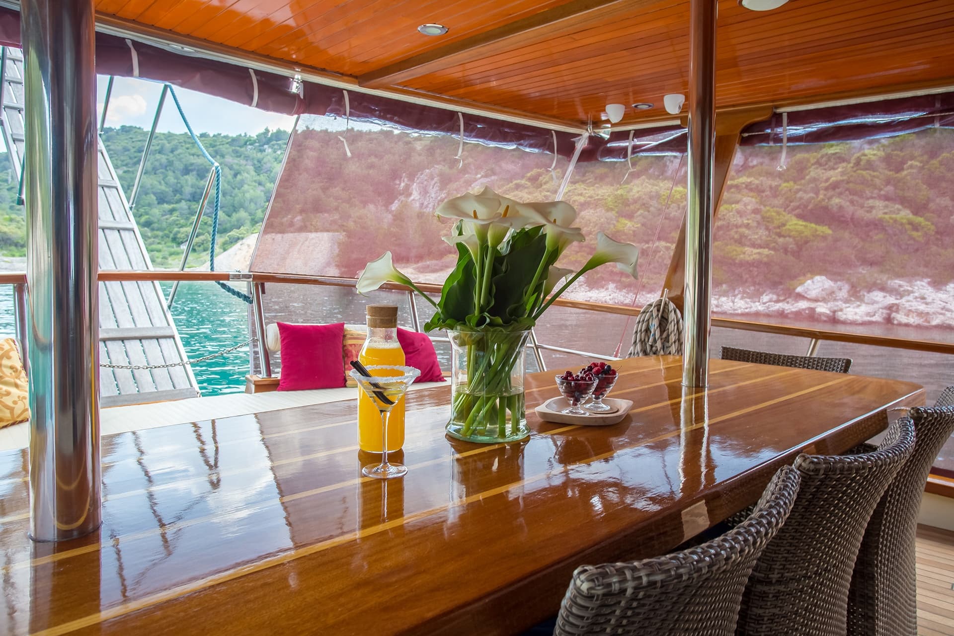 Dining area on boat deck with glossy wood table, flowers, and drinks, overlooking green coastline.