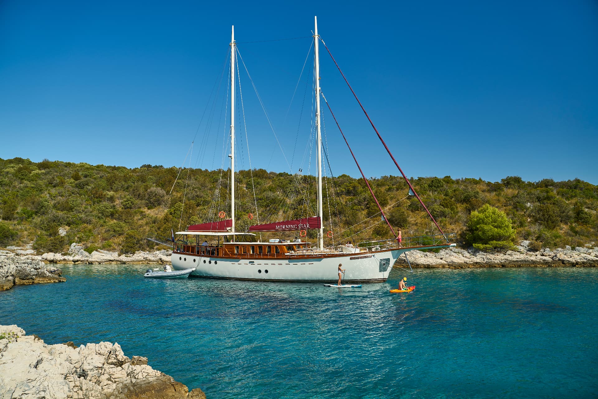 Large sailboat anchored near rocky shore with paddleboarding and kayaking in turquoise water