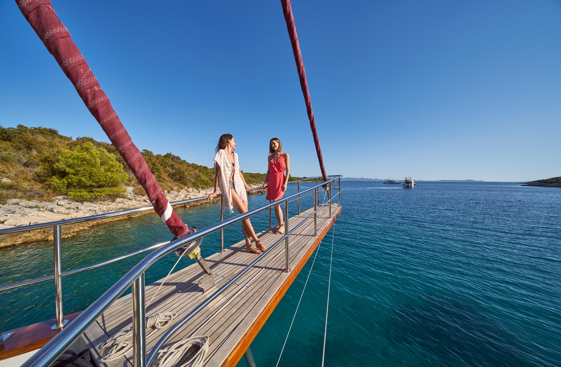 Two women on the bow of a boat near a rocky, green coastline on a sunny day.