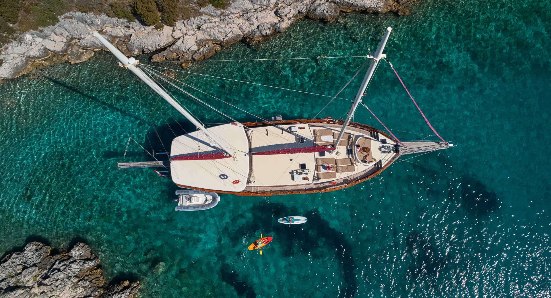 Aerial view of large sailboat anchored near rocky coastline with person kayaking in turquoise water.