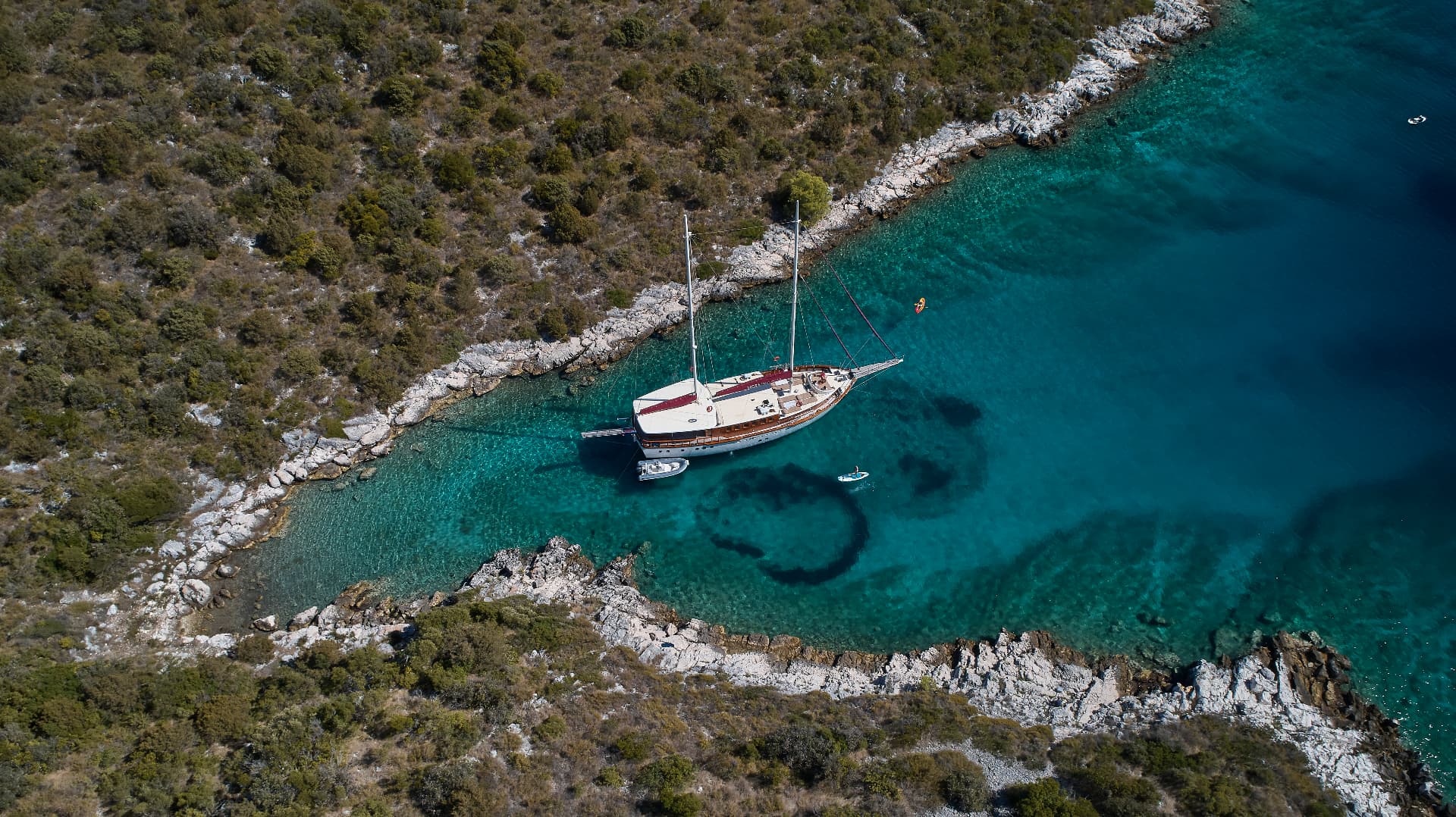Large sailboat anchored in turquoise cove surrounded by rocky, scrub-covered coastline.